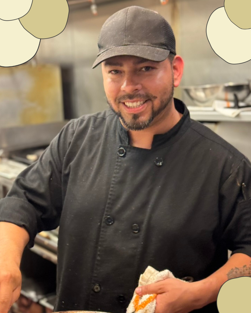 Image of Sous Chef Marco with a baseball cap and black chef attire. he is cooking in the kitchen at the Los Olivos Cafe and has a sweet smile.