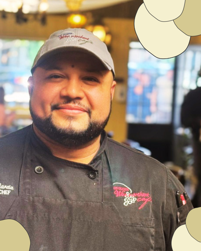 Chef Alex with a beard and go-T wearing a Los Olivos hat and a thoughtful smile inside the Los Olivos Cafe interior with windows in the background