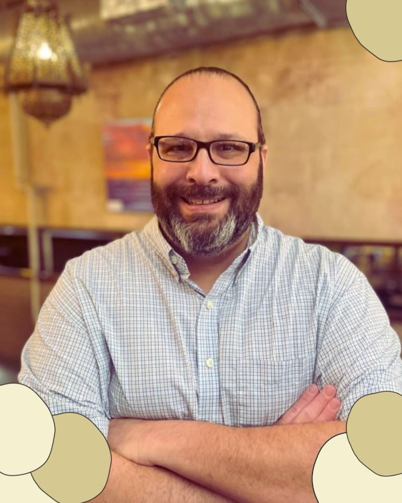 Image of general manager with a smile. He has a beard and black rimmed glasses with his arms crossed in the restaurant's dining room
