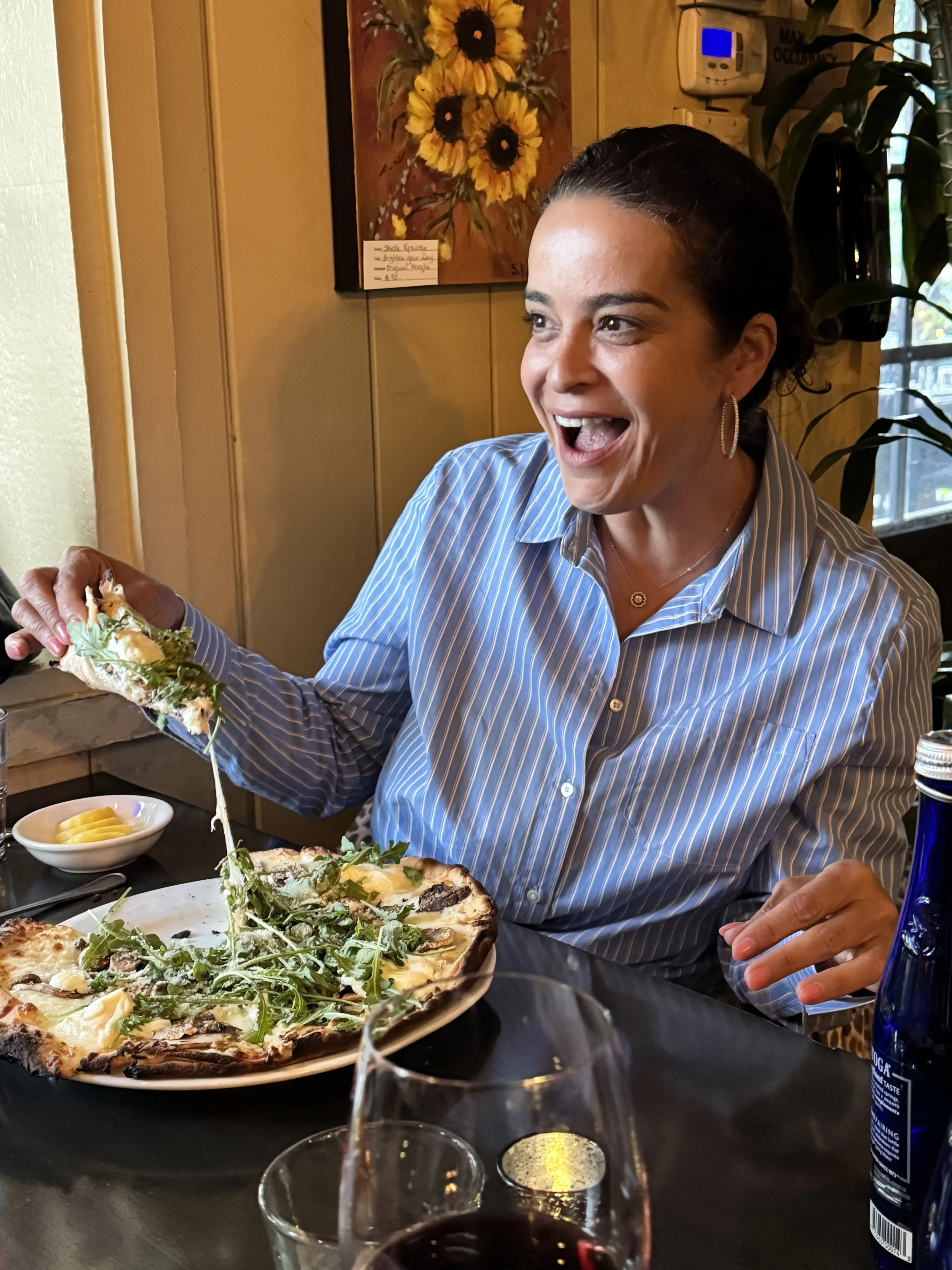 A woman with dark hair and hoop earrings is smiling and holding a slice of Alba pizza while dining at the restaurant. The table has a glass of red wine, and a painting of sunflowers is on the wall behind her.