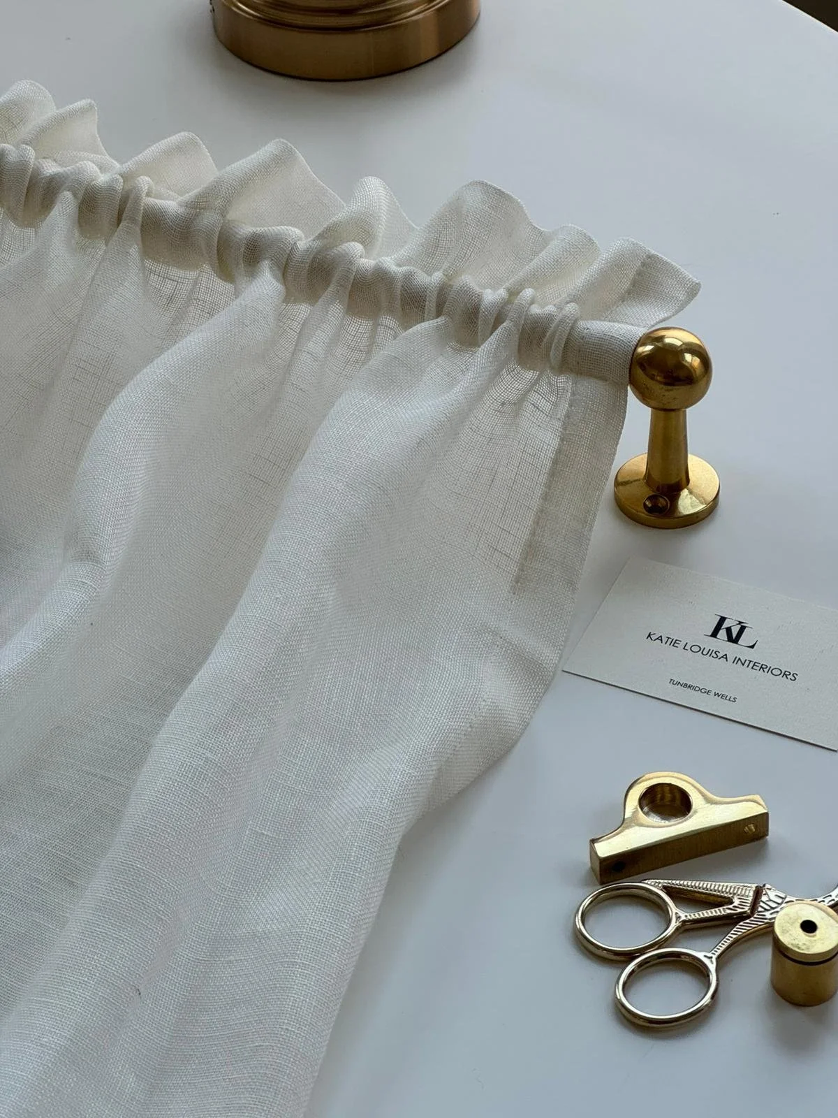 White sheer curtain with a gold curtain rod, scissors, a gold pin, and a business card from Katie Louisa Interiors on a white surface.