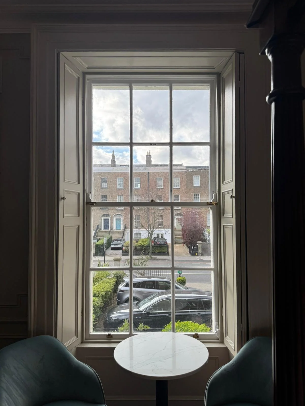A large window with white framing and six panes, overlooking a street with parked cars, bushes, and a brick residential building with steps and hedges, and a partly cloudy sky.
