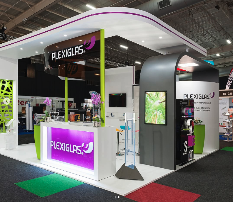 Exhibition booth for PlexiGlas with colorful displays, floral arrangements, and digital screens, set up in a convention center.