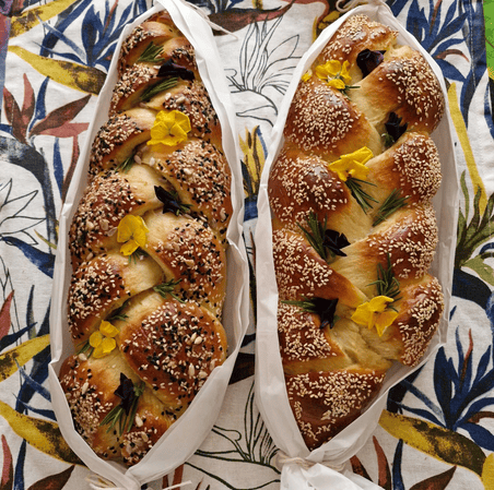 Two braided loaves of bread decorated with sugar, yellow flowers, and green herbs, placed in white paper liners on a patterned cloth.