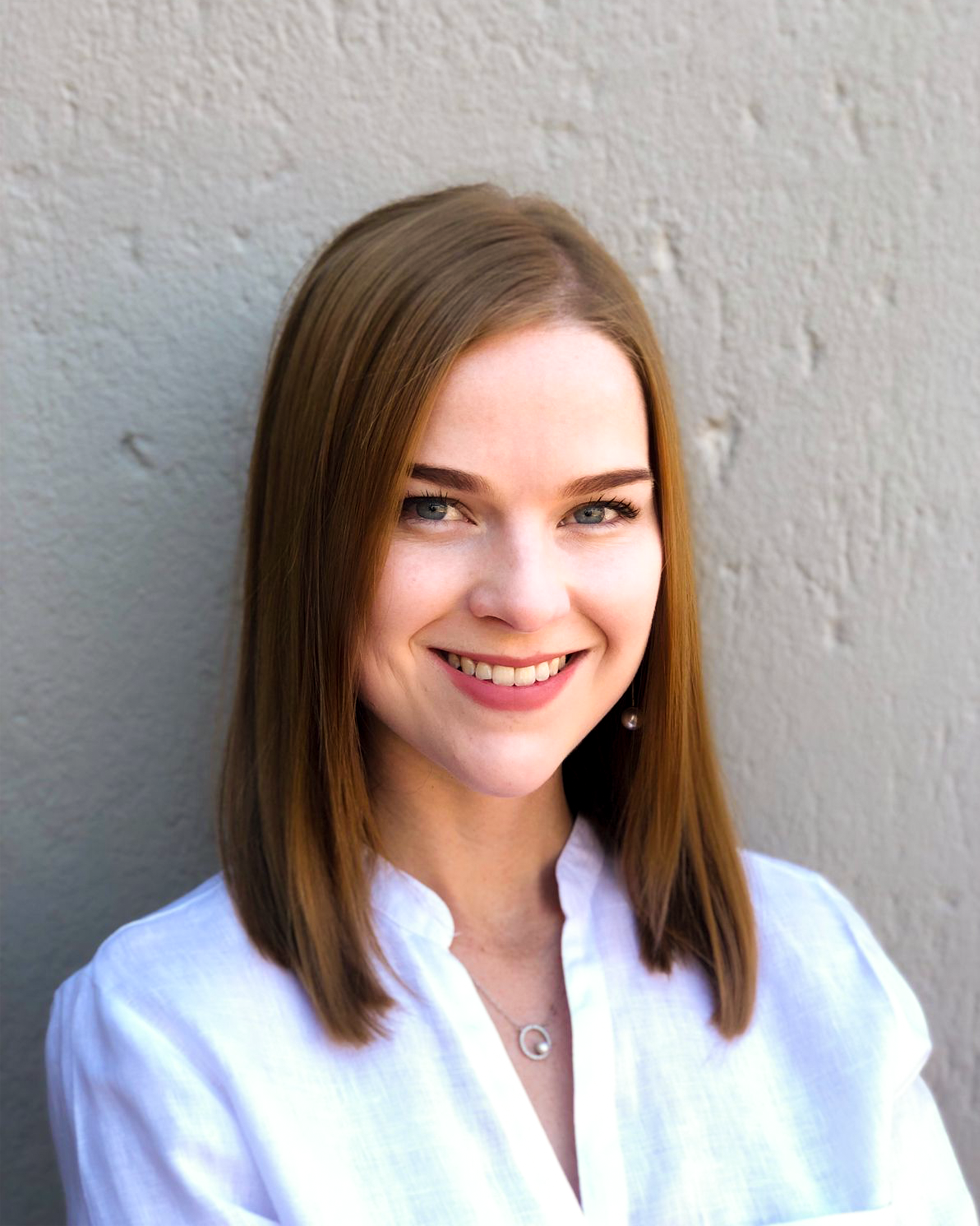 Close-up of a woman's face with straight auburn hair, blue eyes, and a bright smile, wearing a white shirt and a pearl earrings, standing against a textured gray wall.