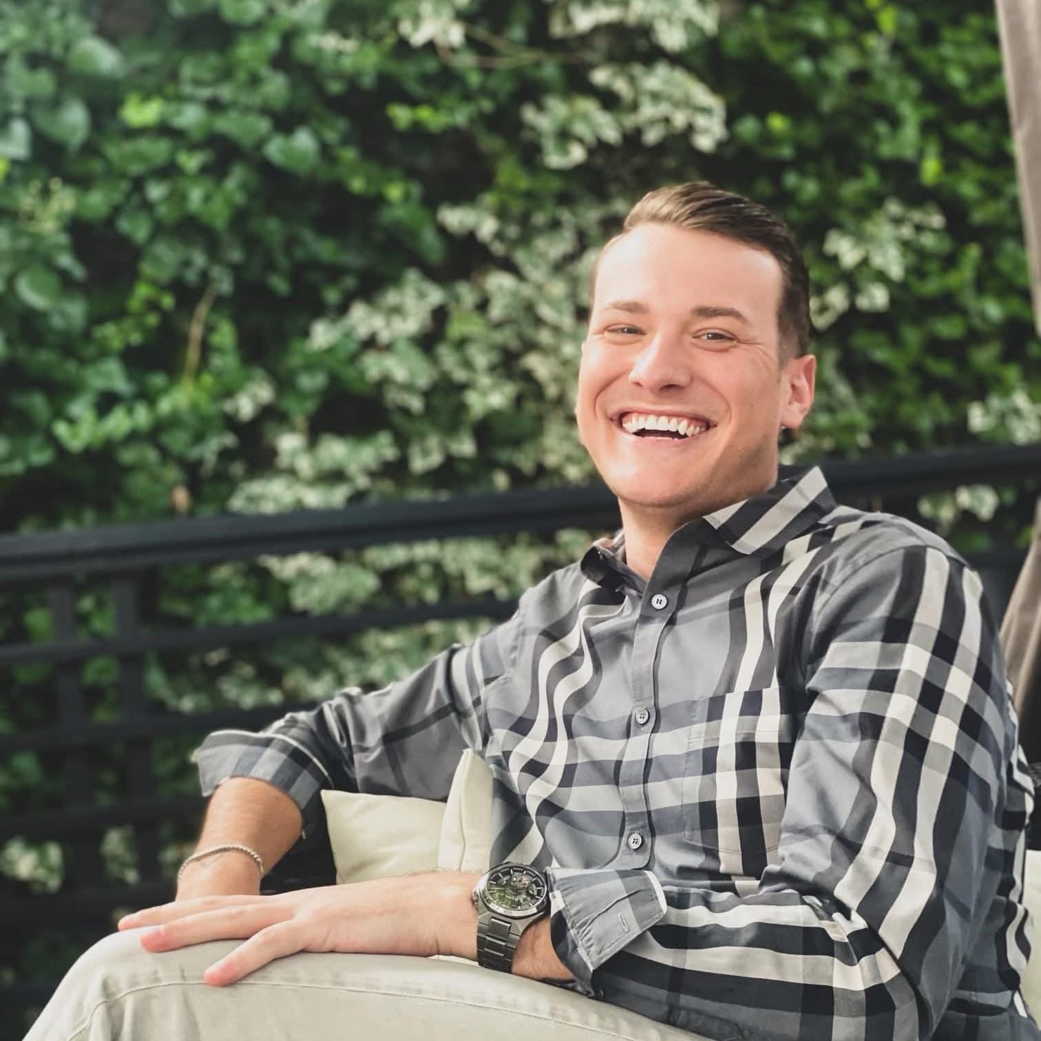A smiling young man sitting outdoors on a cushioned chair, wearing a checkered shirt and a wristwatch, with greenery and white flowers in the background.