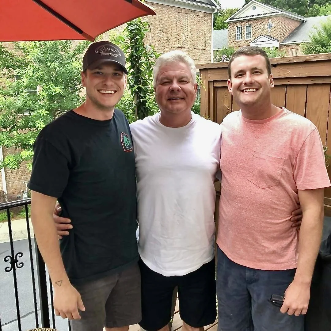 Three men standing together outdoors on a patio, smiling at the camera. The man on the left is wearing a black T-shirt and a black cap, the man in the middle is in a white T-shirt, and the man on the right is in a pink T-shirt. There is a red umbrella above them, a wooden fence behind, and greenery and houses in the background.