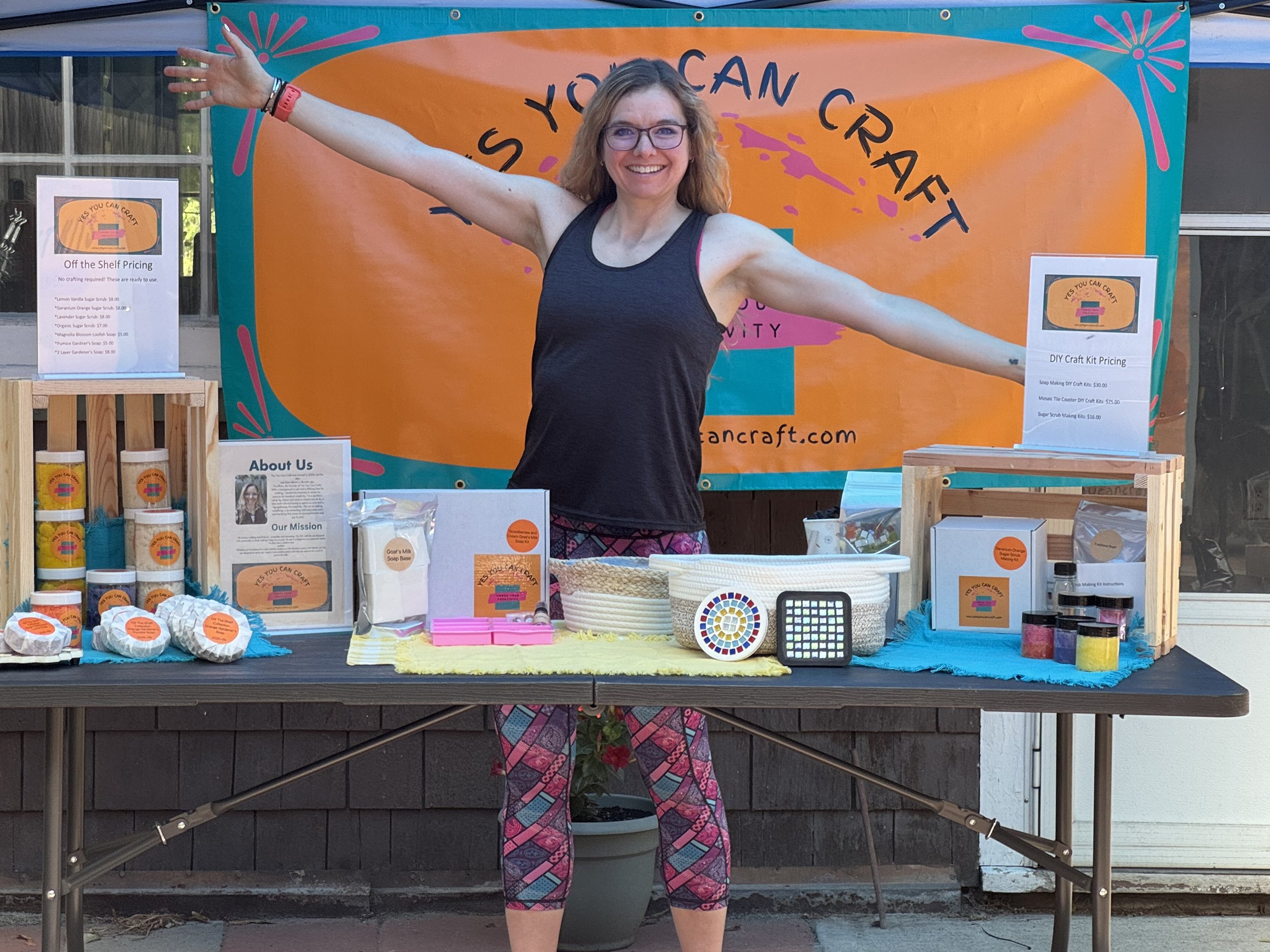 Women standing behind a craft table at a craft fair booth with a large orange and teal banner that reads "Serious You Can Craft." The table displays soap, candles, and craft kits for sale. There is an "About Us" sign and pricing signs for craft kits.