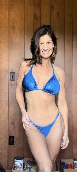 Woman with shoulder-length dark hair wearing a blue bikini standing in front of a wooden wall.