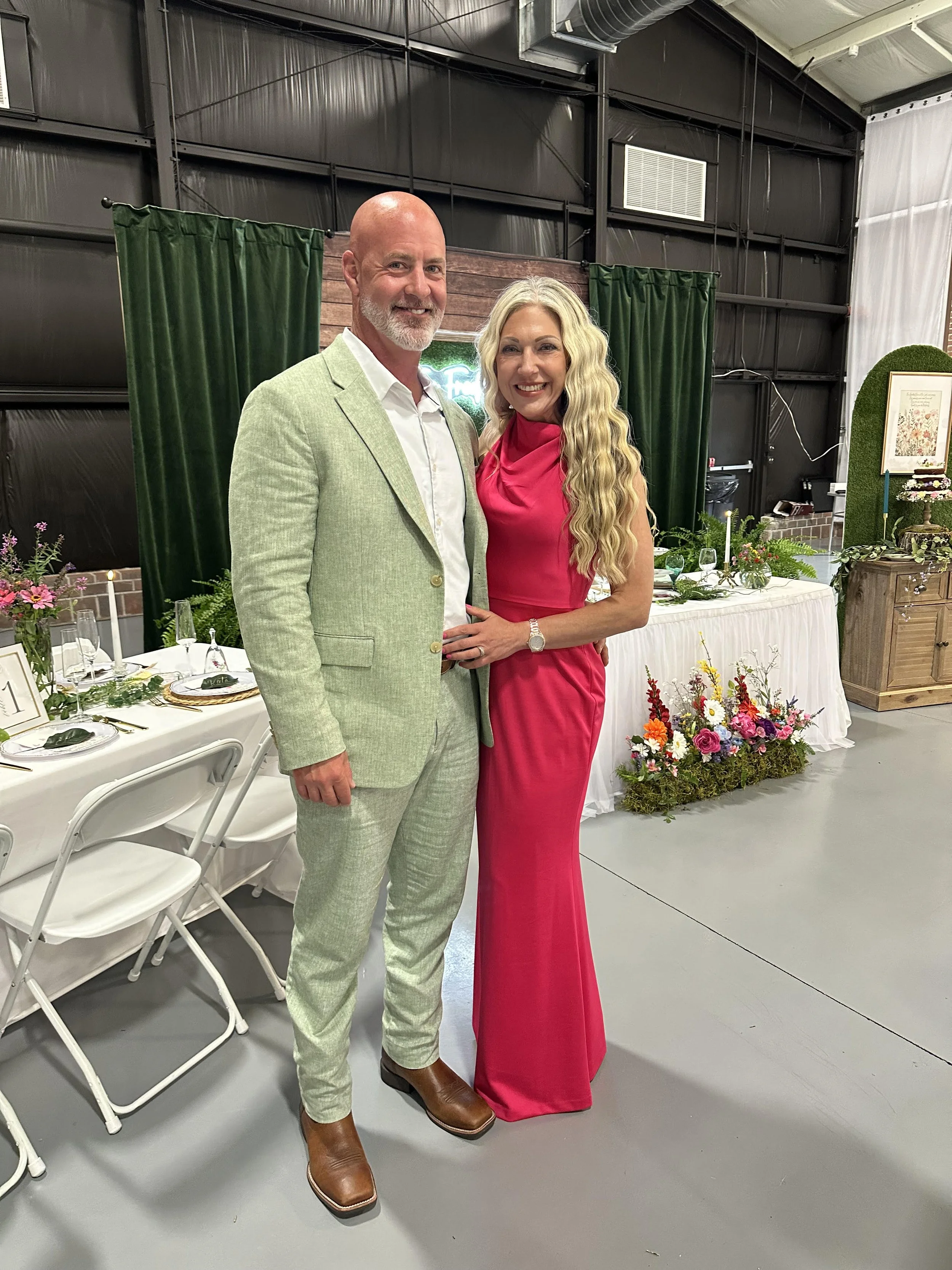 A man in a light green suit and a woman in a bright pink dress standing together and smiling at an indoor event with decorated tables and floral arrangements.