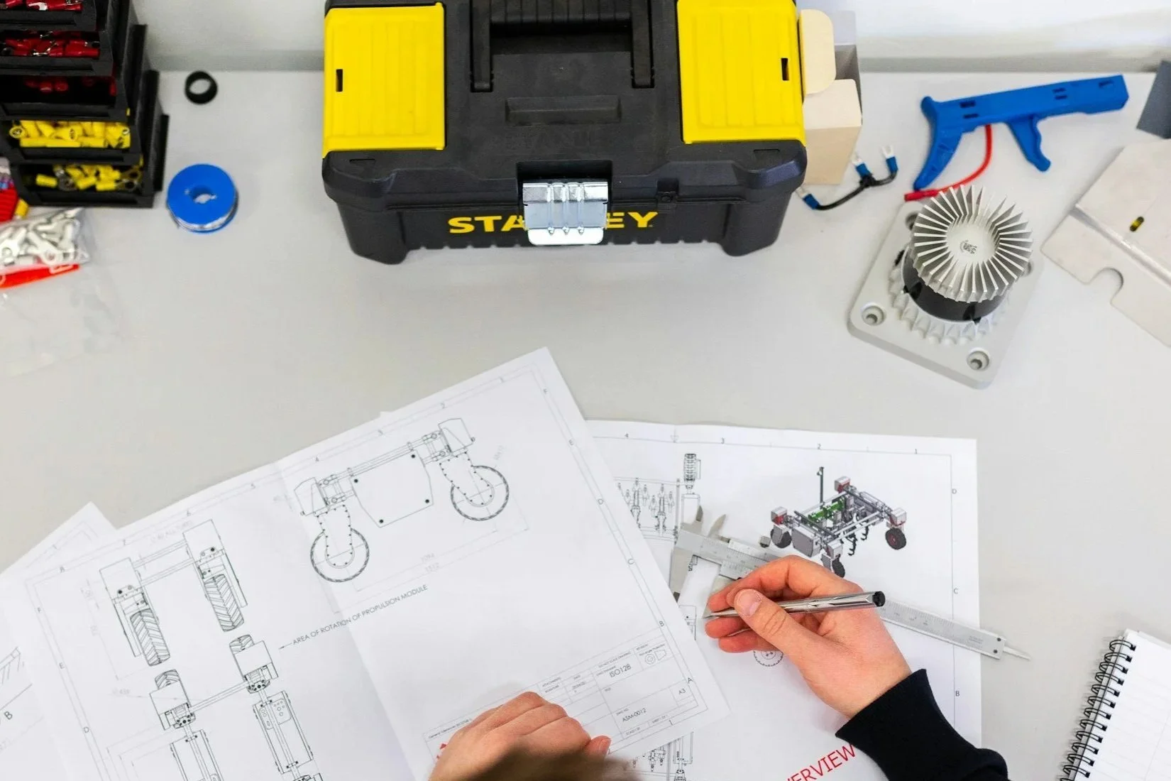 Technical drawings and schematics of a robotic or mechanical system on a table, with a person's hands measuring with a caliper, organized tools, and a Stanley toolbox in the background.