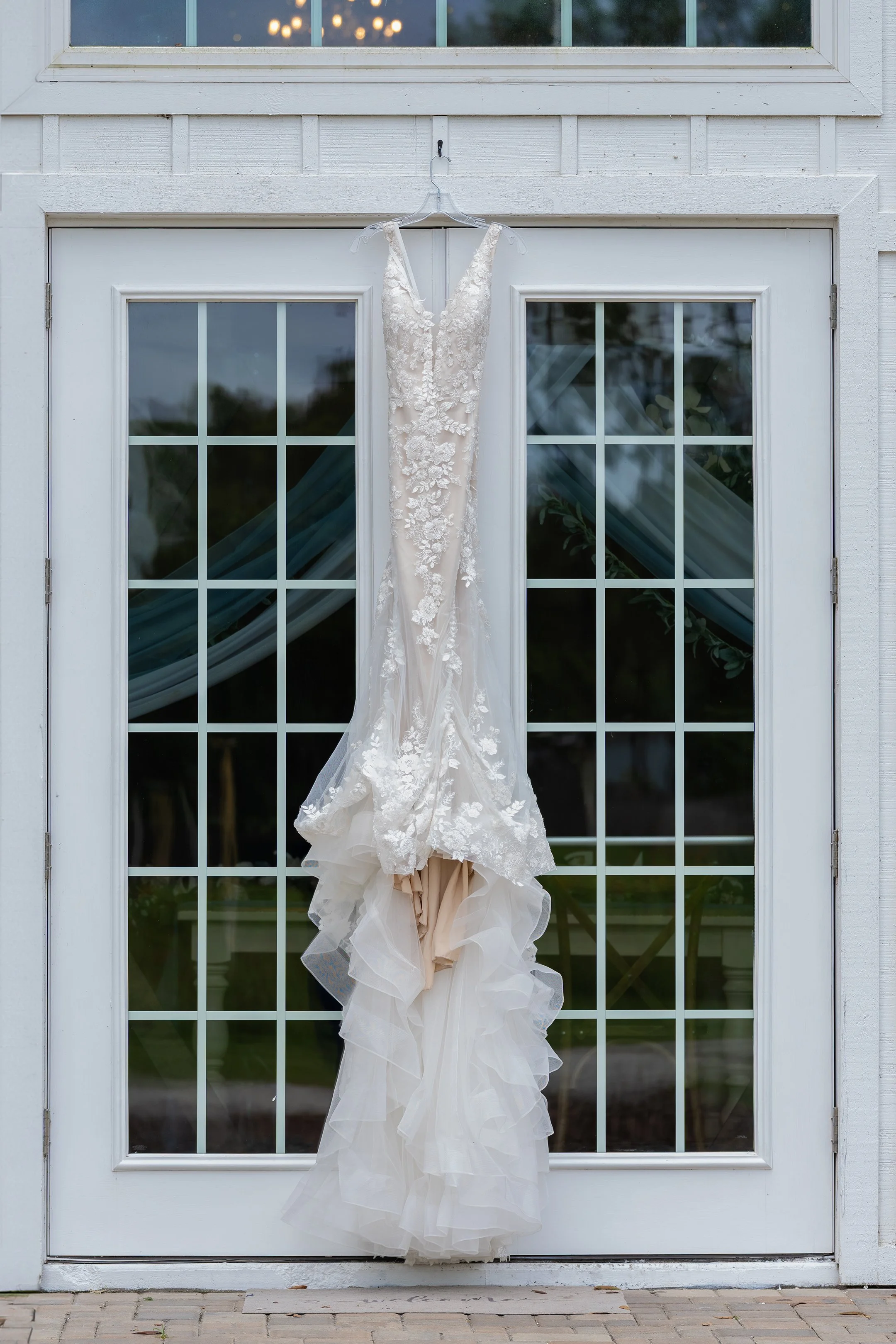 A wedding dress hanging on a hanger outside a white house with glass doors, under a window, with curtains and greenery visible inside.