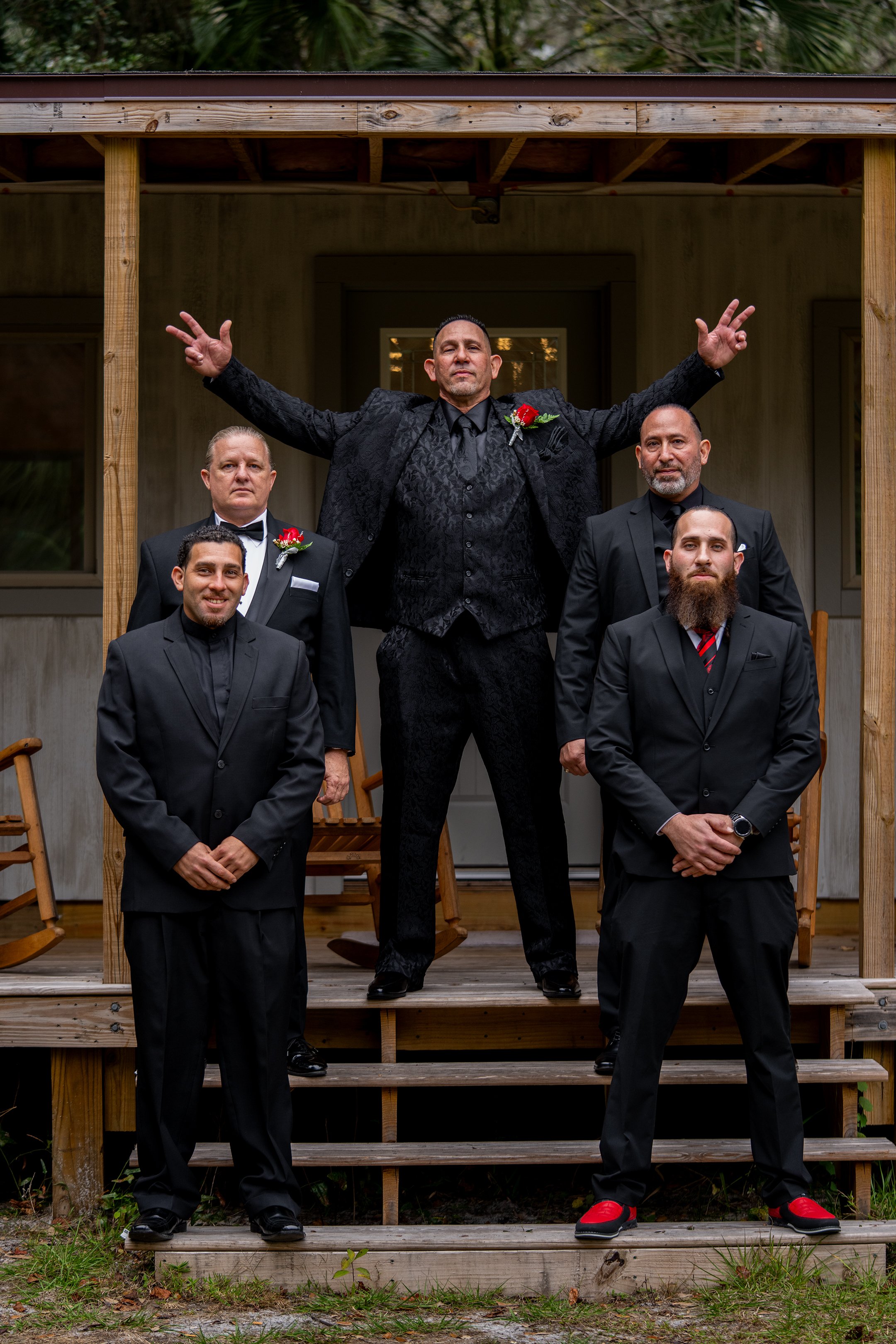 Five men in suits standing on the steps of a porch, with one man in the middle raising his arms, wearing a black suit with a red boutonniere, surrounded by four other men in black suits, two with red boutonnieres and two without.