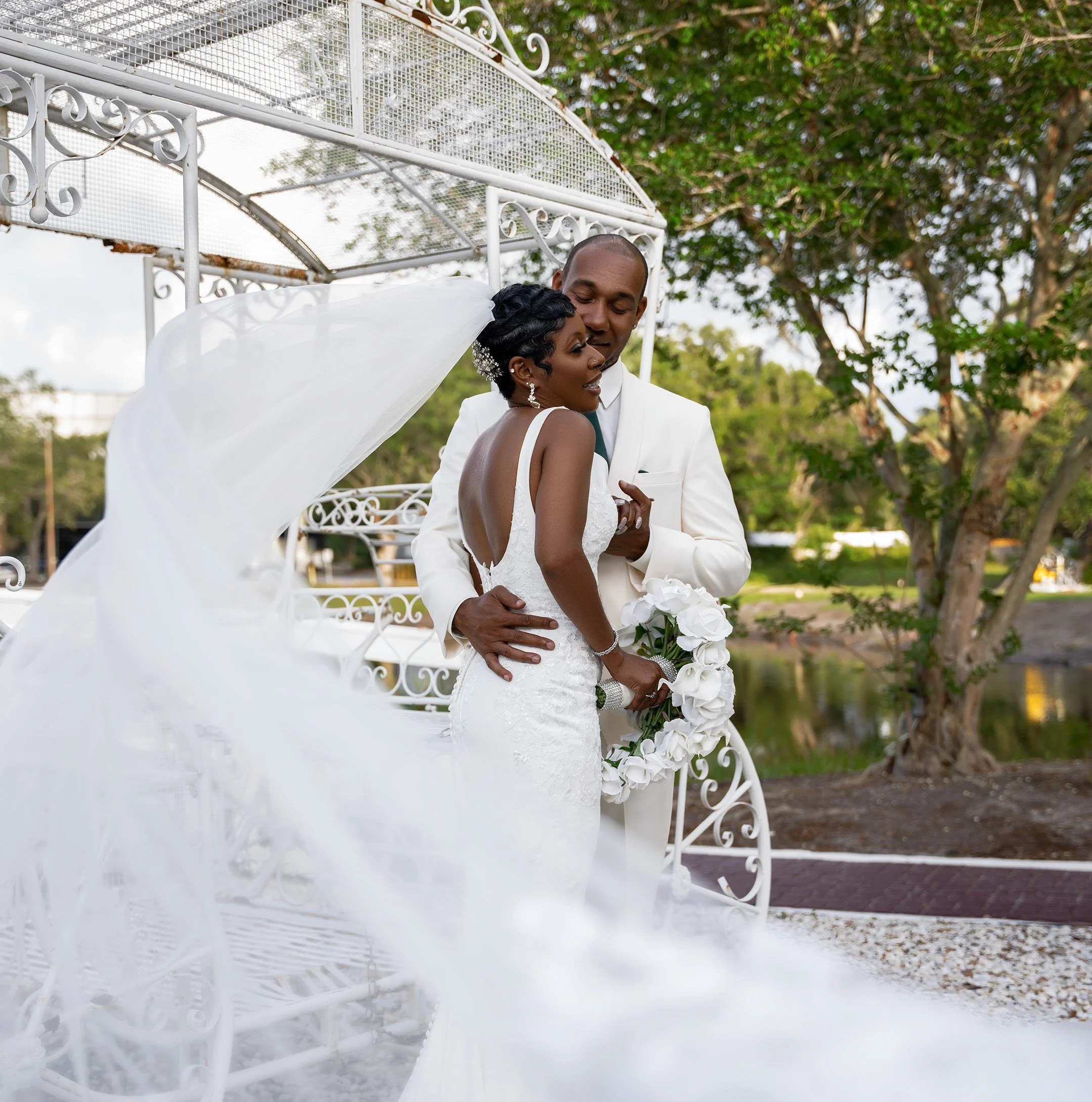 A newlywed couple embracing outdoors near a white decorative carriage, with green trees and a pond in the background. The bride is wearing a white lace wedding dress and veil, holding a bouquet of white roses. The groom is dressed in a white suit.