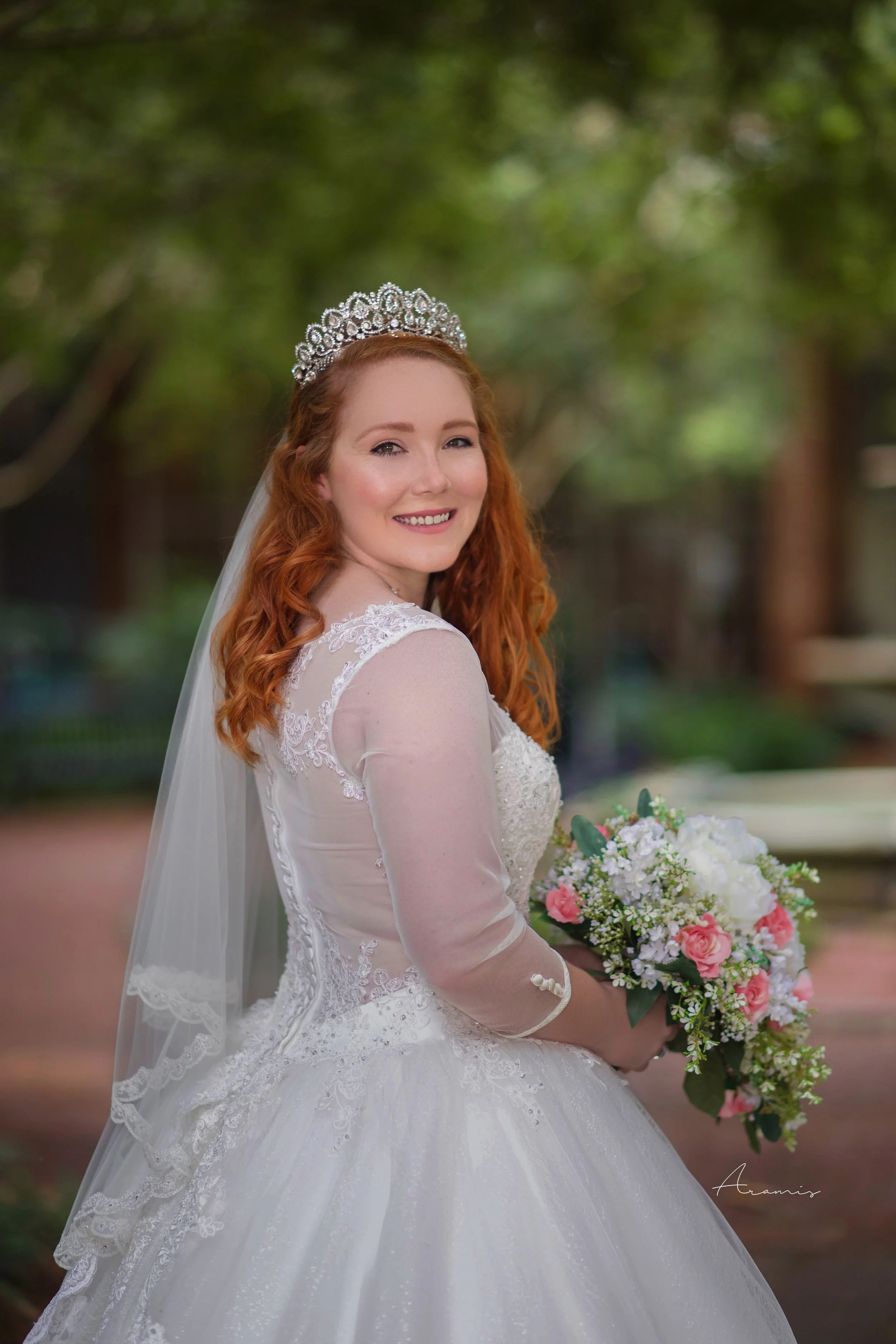 A bride with red hair wearing a wedding dress, tiara, and veil holds a bouquet of pink and white flowers, outdoors with a blurred green background.