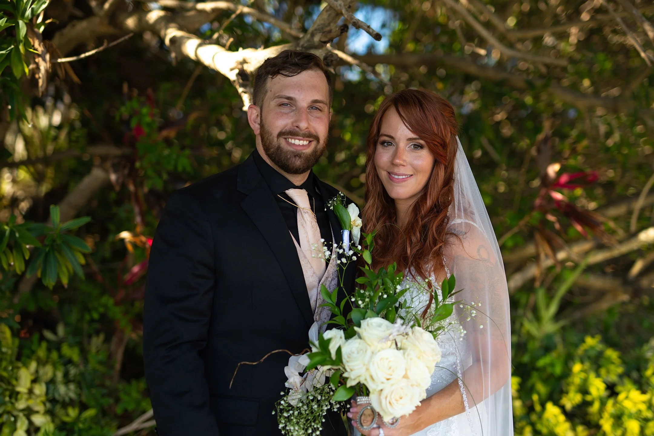 A newlywed couple smiling outdoors, standing close together, with the groom in a black suit and the bride in a white wedding dress holding a bouquet of white roses and greenery.