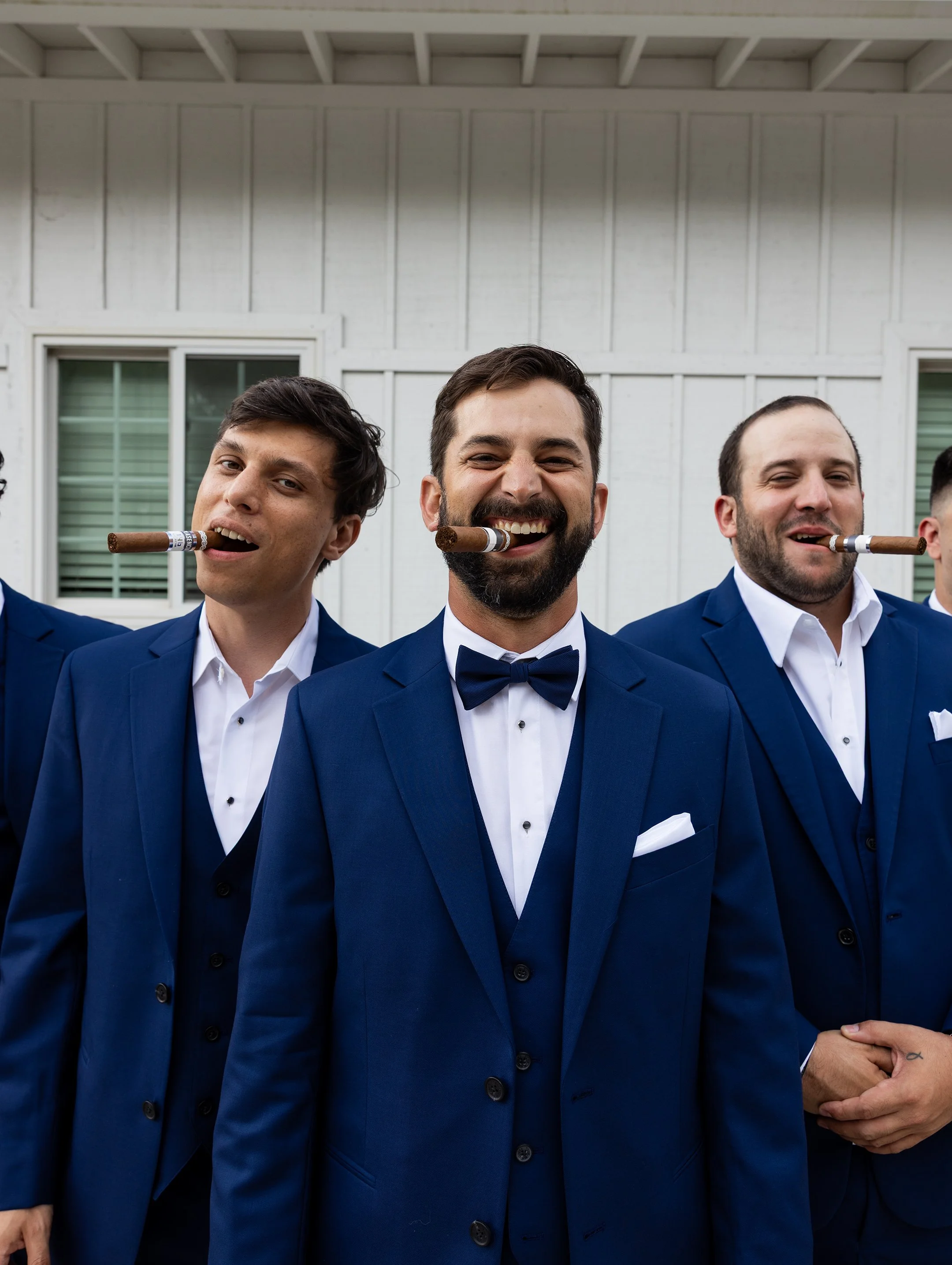 Group of men in blue tuxedos and white shirts standing outside, holding cigars in their mouths, smiling.