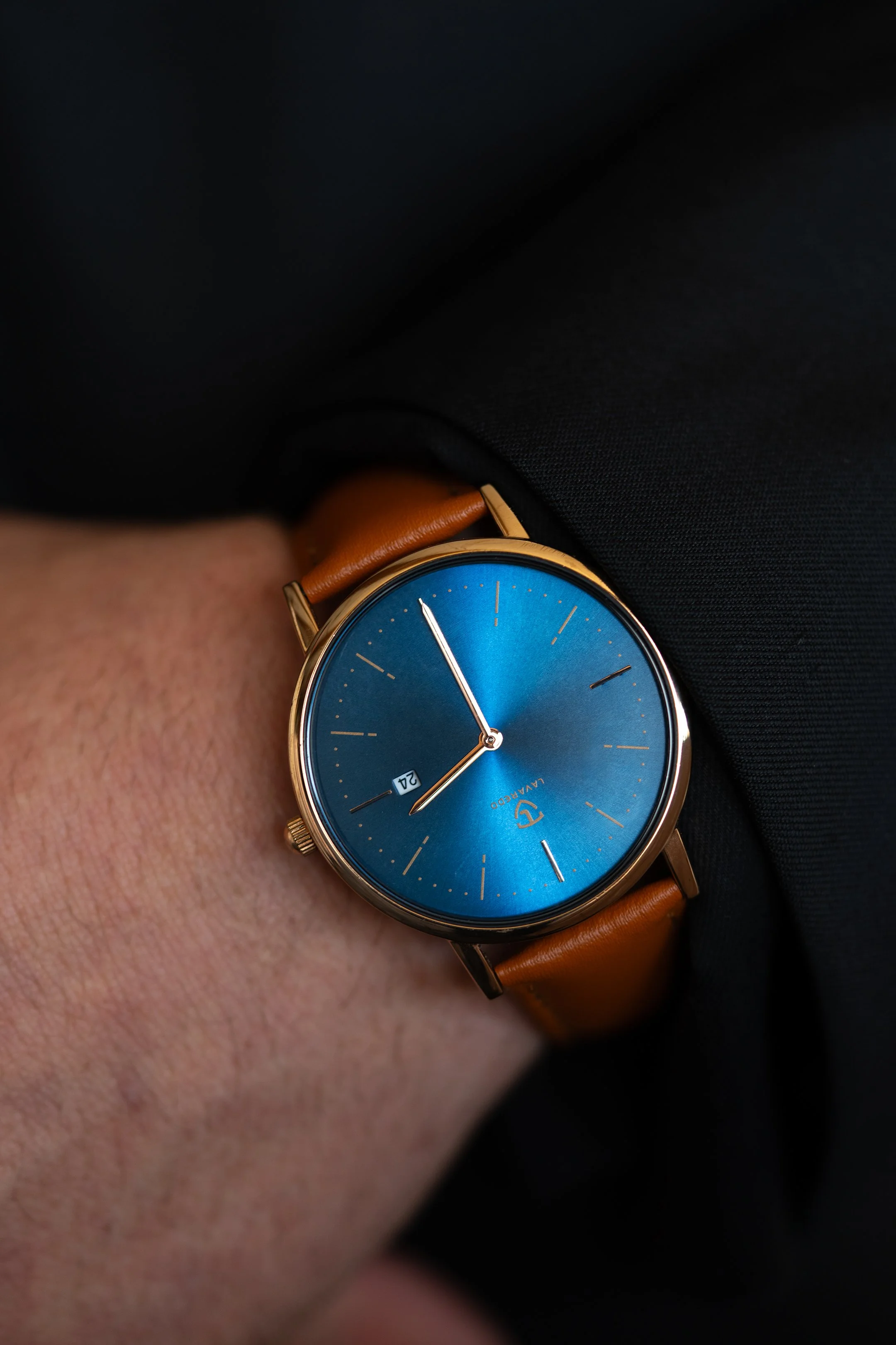 A person's wrist wearing a gold-tone watch with a blue dial and brown leather strap, showing the time as 8:55 and the date as the 21st, against a black fabric background.