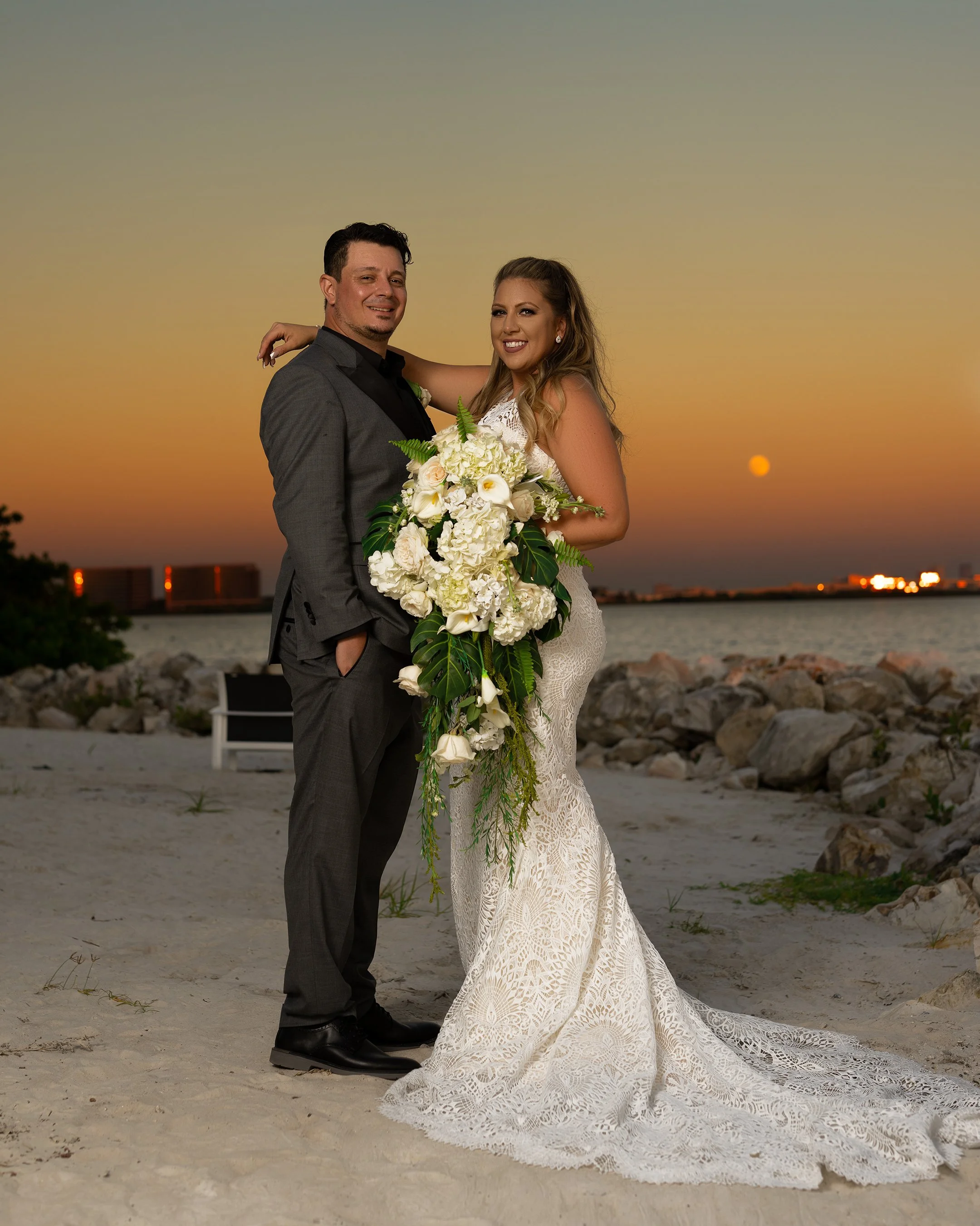 Bride and groom standing on beach at sunset, holding a large bouquet of white flowers, with the city skyline and water in the background.