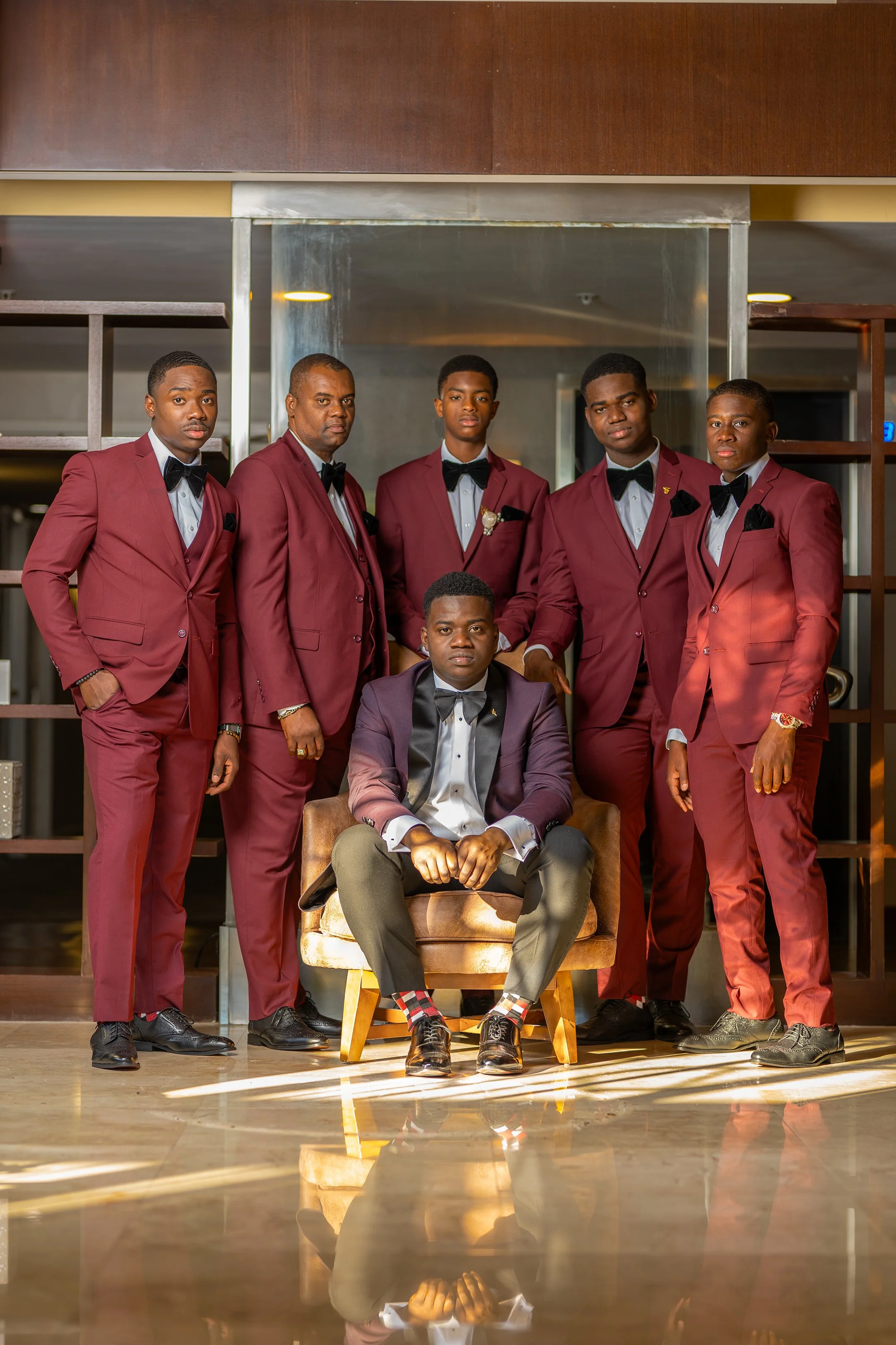 Six men dressed in formal suits, five standing and one seated in front, posing for a photo indoors.