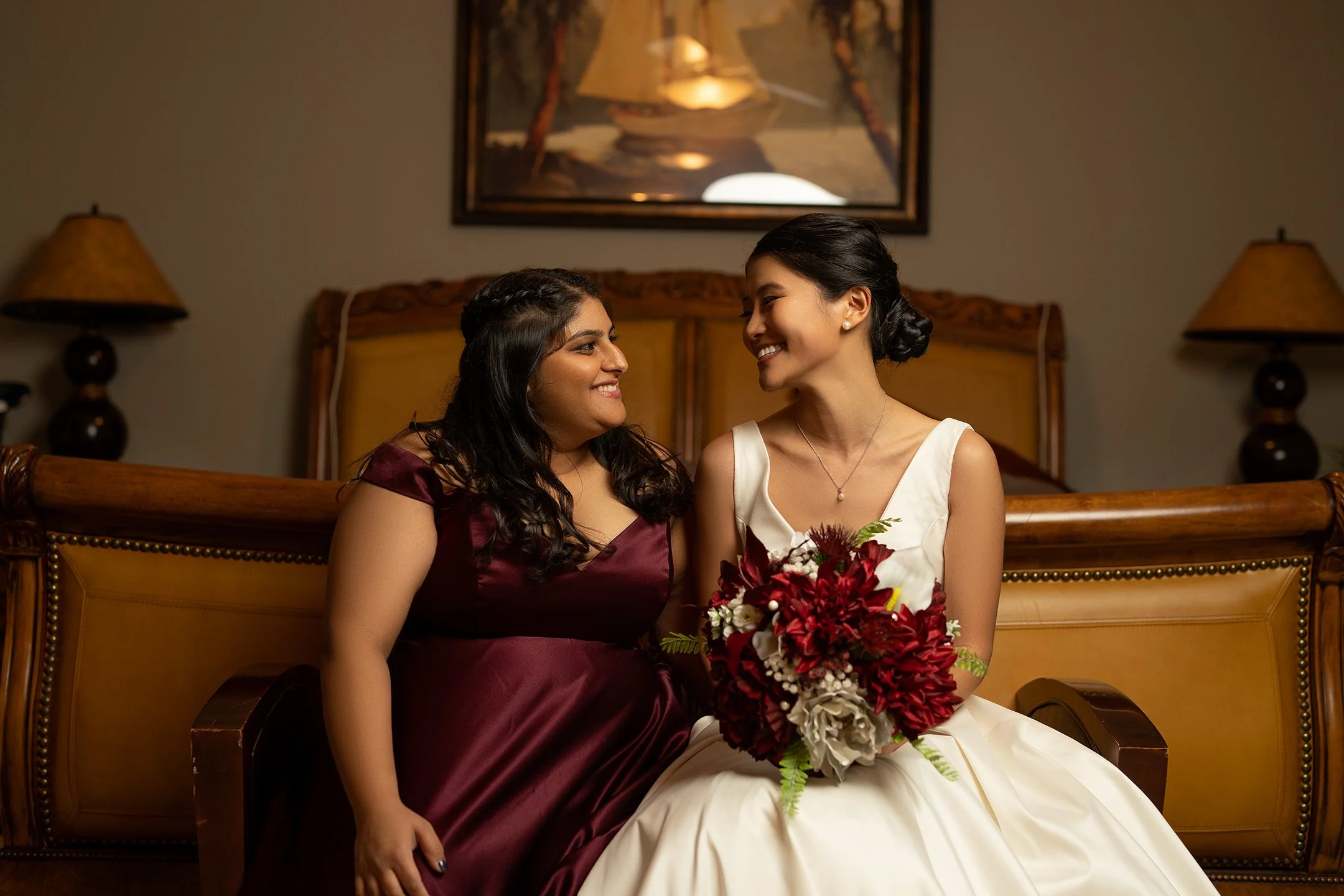 Two women sitting on a vintage wooden sofa, one in a white wedding dress holding a bouquet of red and white flowers, smiling at each other, in a warmly lit room with a painting and lamps in the background.