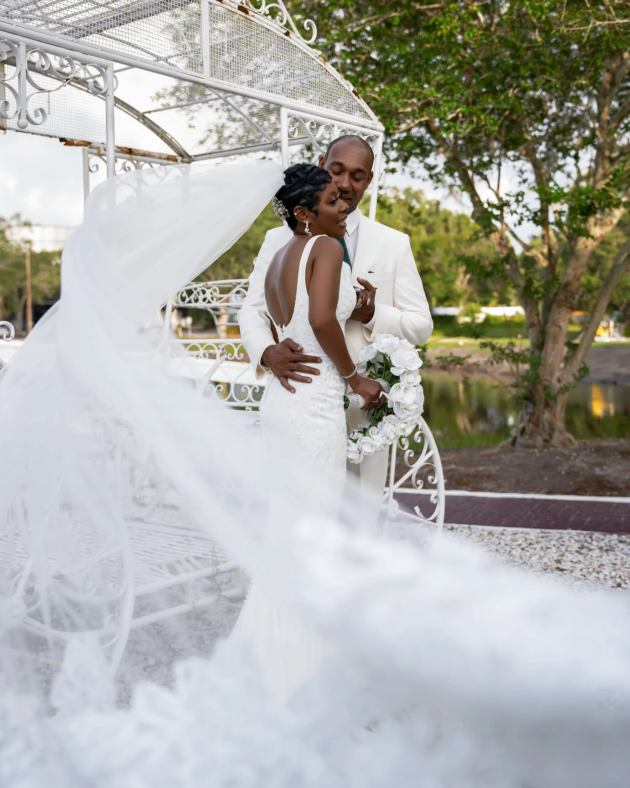 A bride and groom embrace outdoors, with a white ornate cart and trees in the background, during sunset. The bride wears a white lace wedding gown with a flowing veil, and the groom wears a white tuxedo. The bride holds a white floral wreath.
