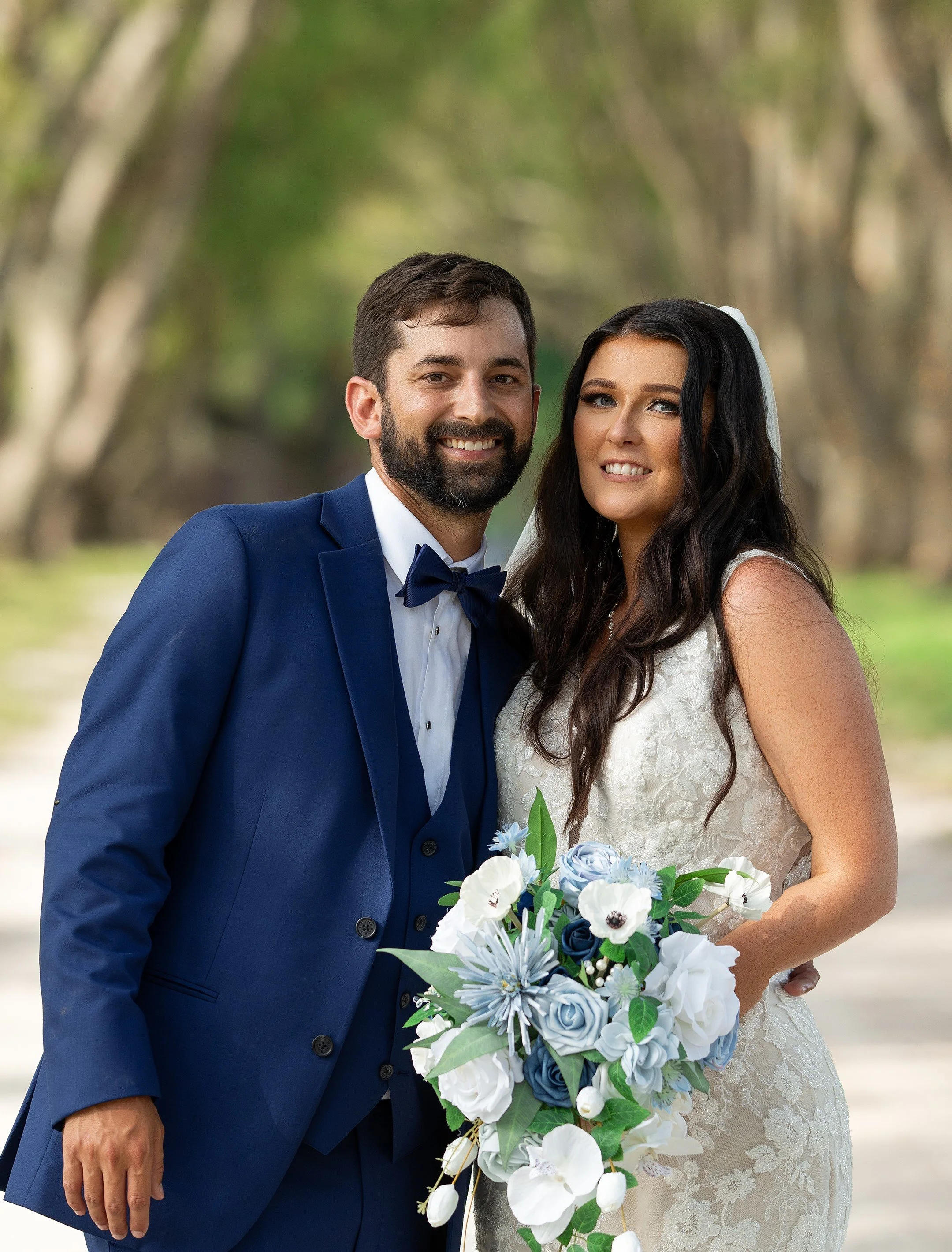 A newlywed couple outdoors, smiling at the camera. The groom wears a blue suit with a bow tie, and the bride wears a lace wedding dress holding a bouquet of blue, white, and green flowers with a white veil in her dark, wavy hair.