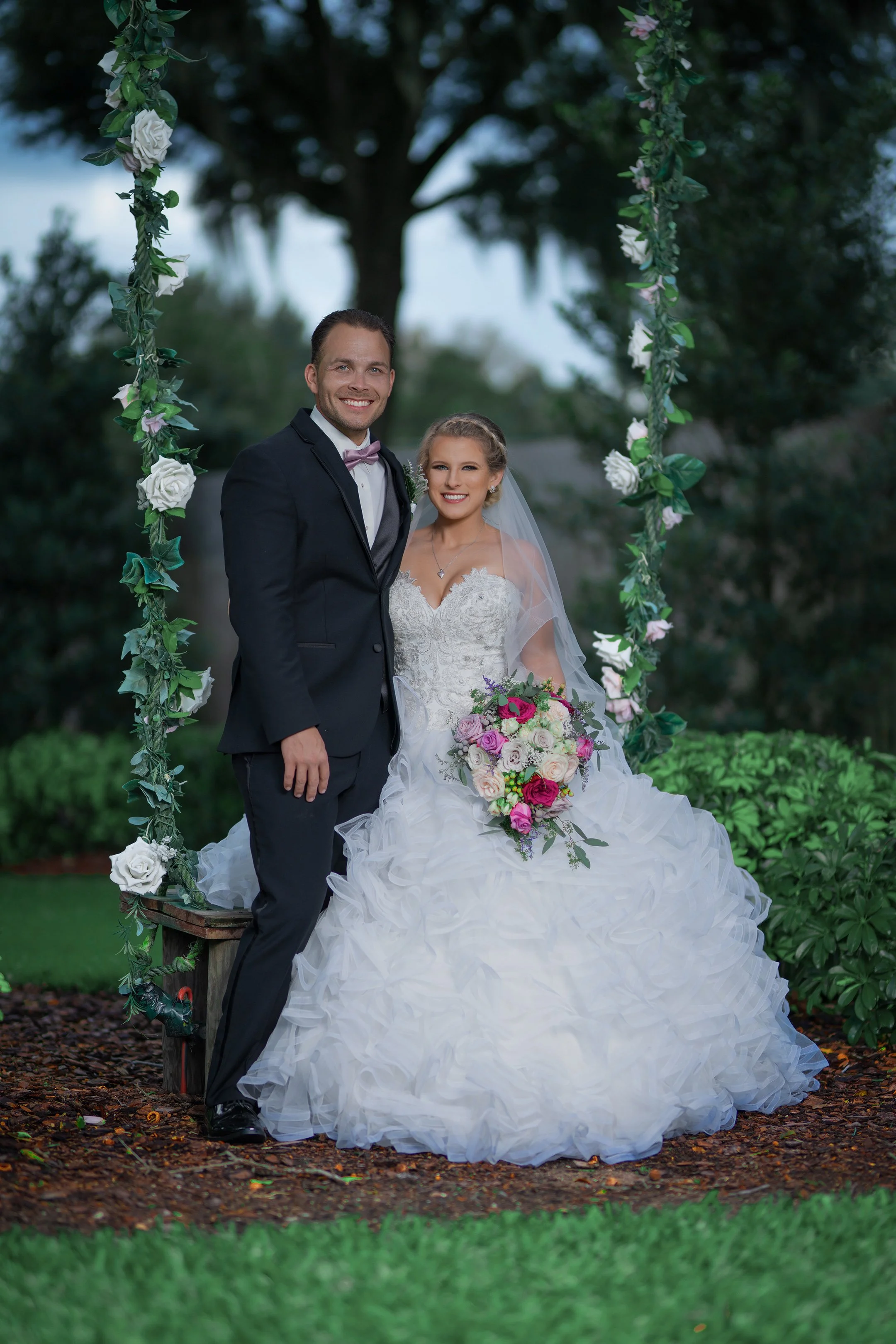 A bride and groom in wedding attire posing on a decorated outdoor arbor with flowers, greenery, and a large tree in the background.