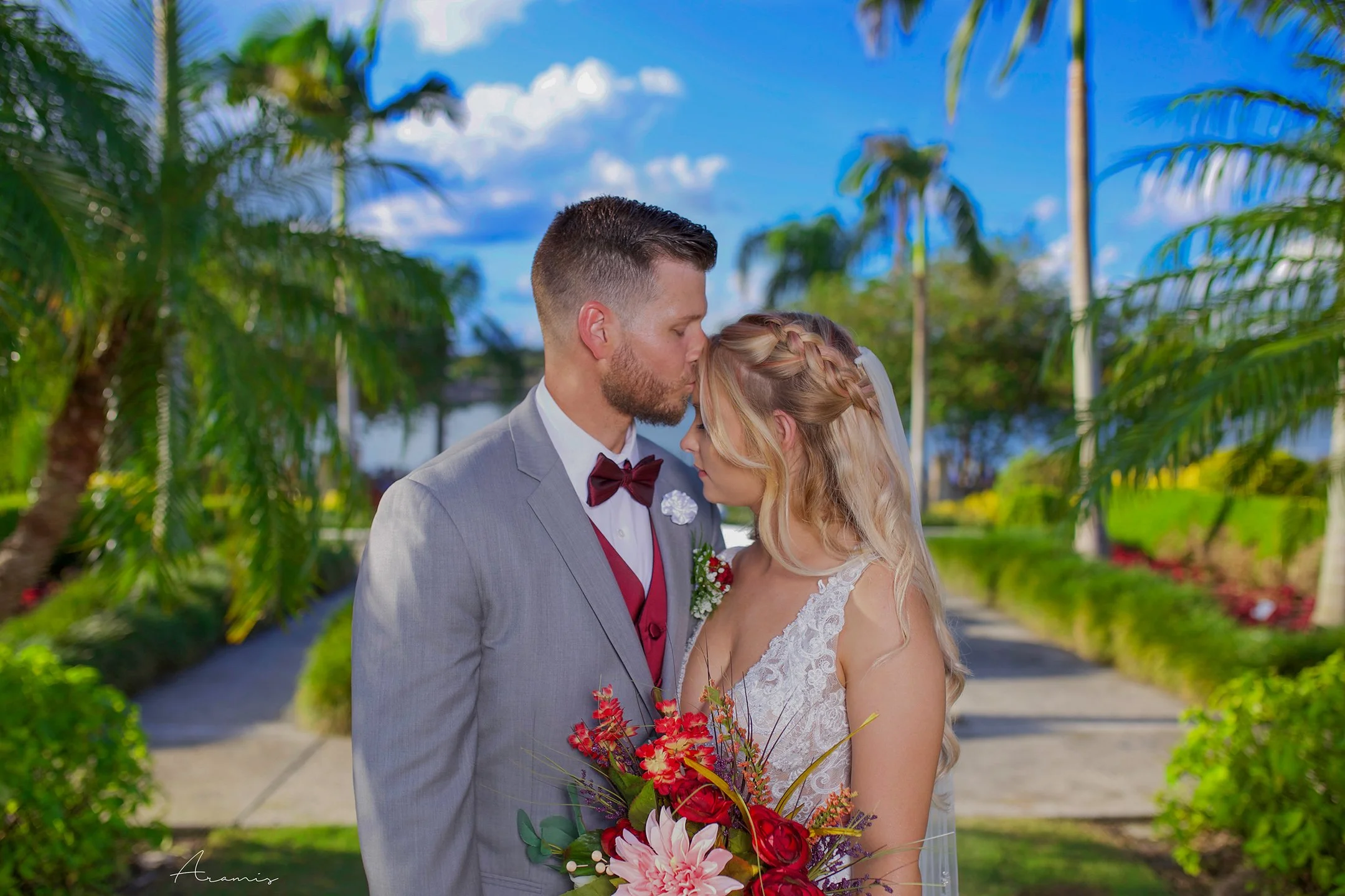 A bride and groom standing close outdoors on a sunny day, with palms and greenery in the background. The groom is kissing the bride on her forehead, and they are holding a bouquet of red and pink flowers.