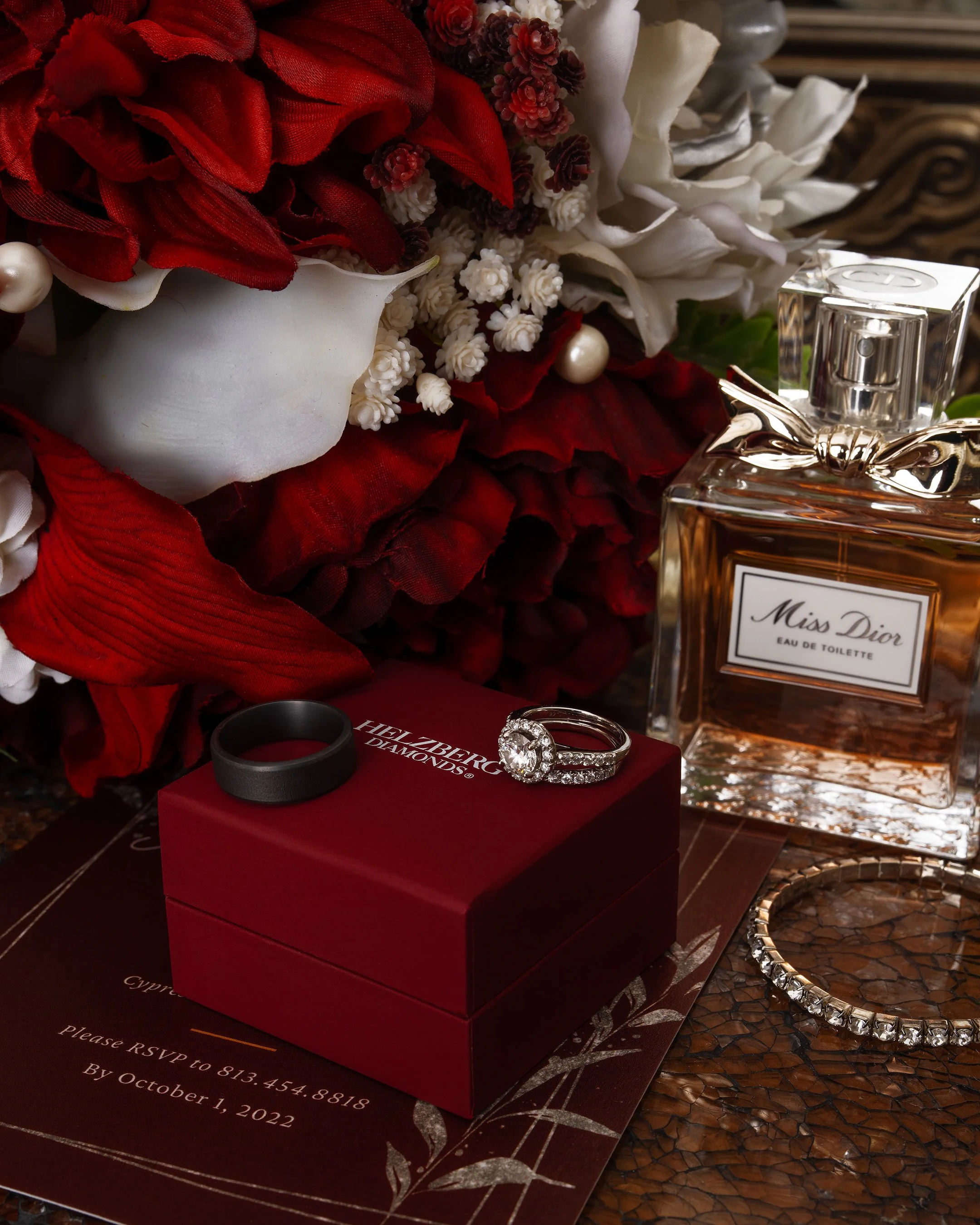 A wedding ring set on a red jewelry box with a smaller black band, surrounded by red and white flowers, a perfume bottle labeled Miss Dior, and a diamond bracelet, all on a decorative surface.