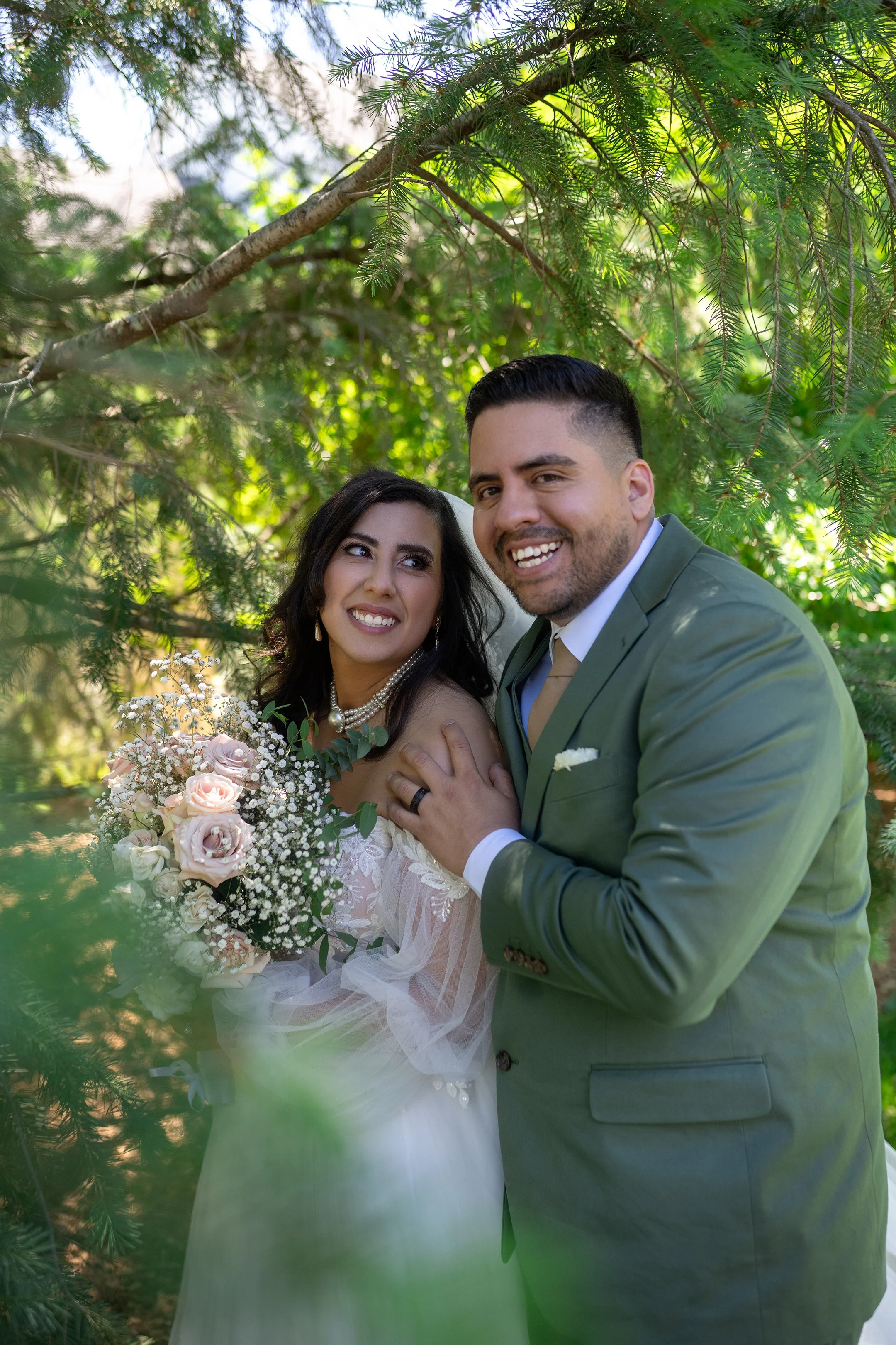 A newlywed couple standing among green trees, the bride holding a bouquet of pink and white roses, smiling and wearing a white wedding dress with lace details, and the groom wearing a green suit, white shirt, and beige tie, both smiling.
