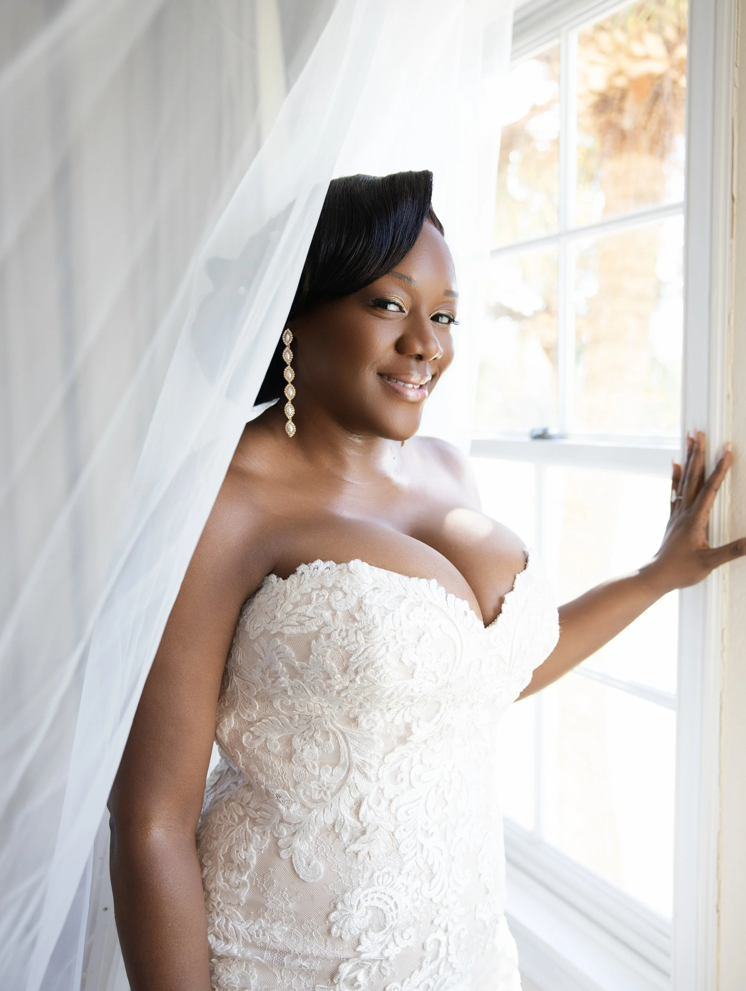 A woman in a wedding dress standing by a window, smiling, with a veil partially covering her head.
