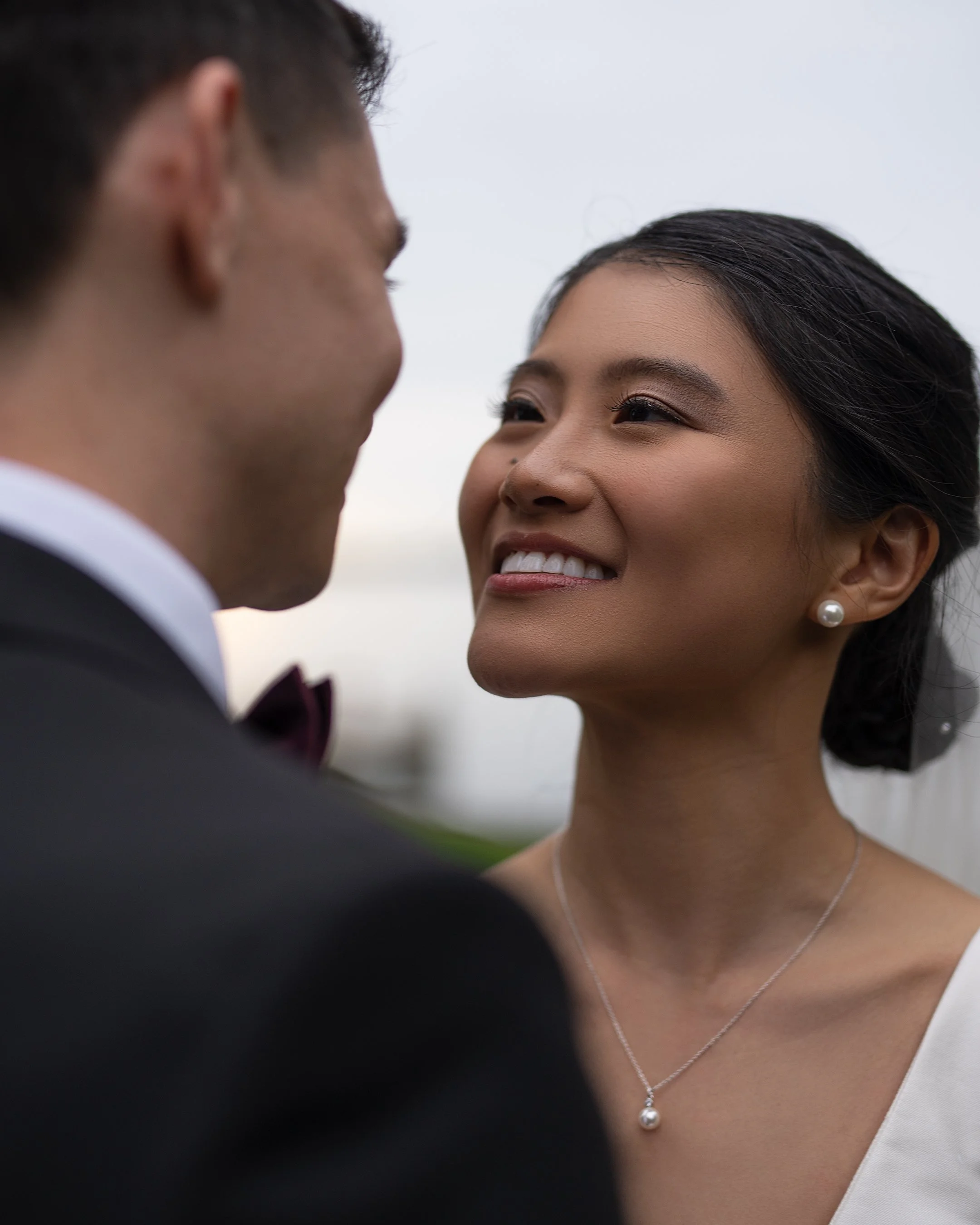 Close-up of a happy woman with pearl earrings and necklace, smiling at a man in a tuxedo during a wedding or romantic moment.