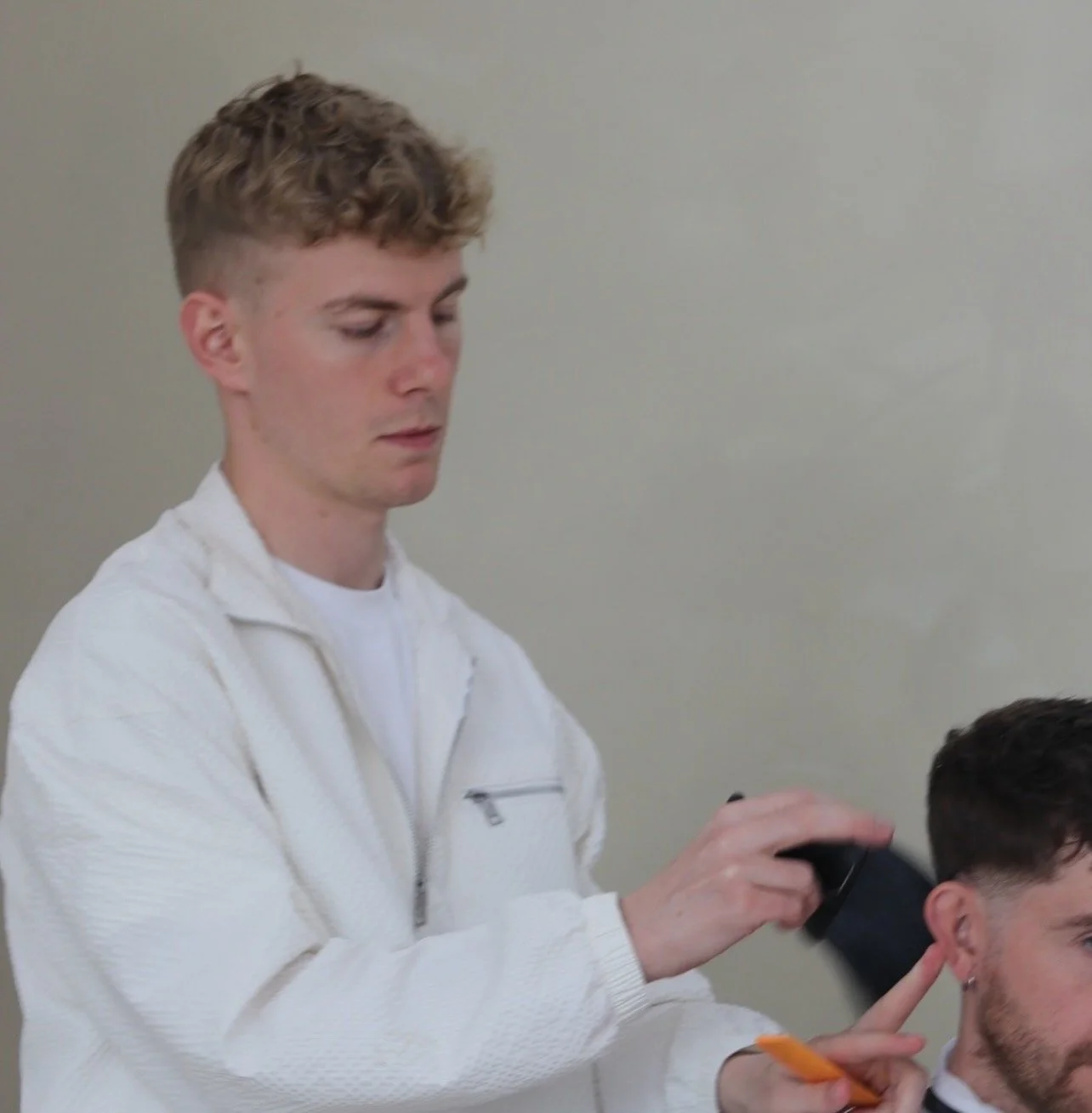Founder Rohin O'Neill cuts another person's dark brown hair in Column barbershop. The person receiving the haircut is seated, while the Rohin stands behind him, carefully trimming his hair with clippers. The background is plain, grey limewash wall