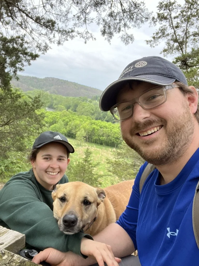 Selfie of a man (John), woman (Christina), and dog (Bailey) with a background overlooking a natural area