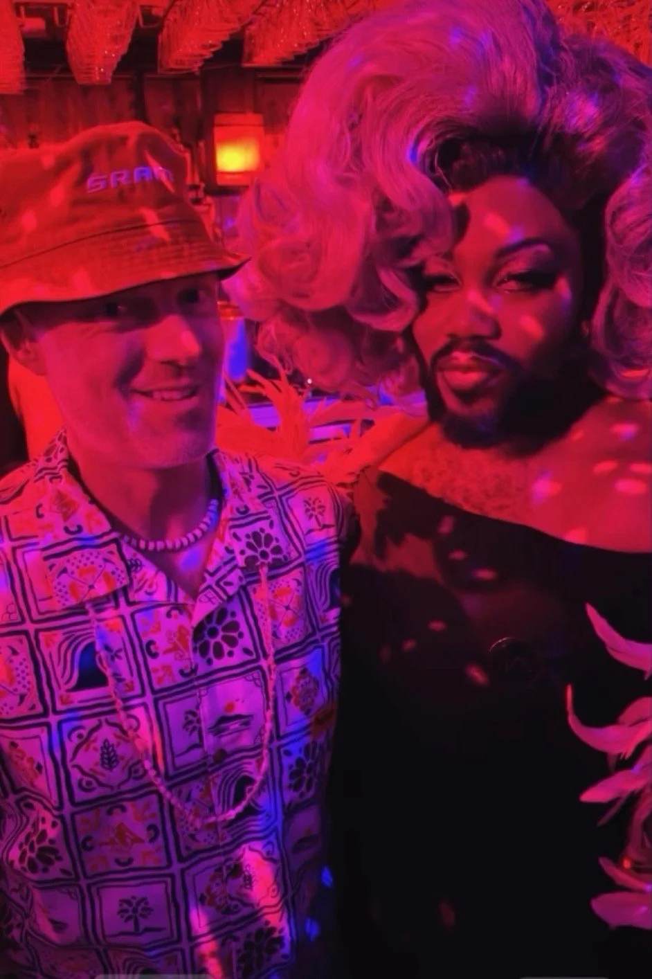 man in red bucket hat next to man with pretty face and blonde wavy wig in a nightlife setting.