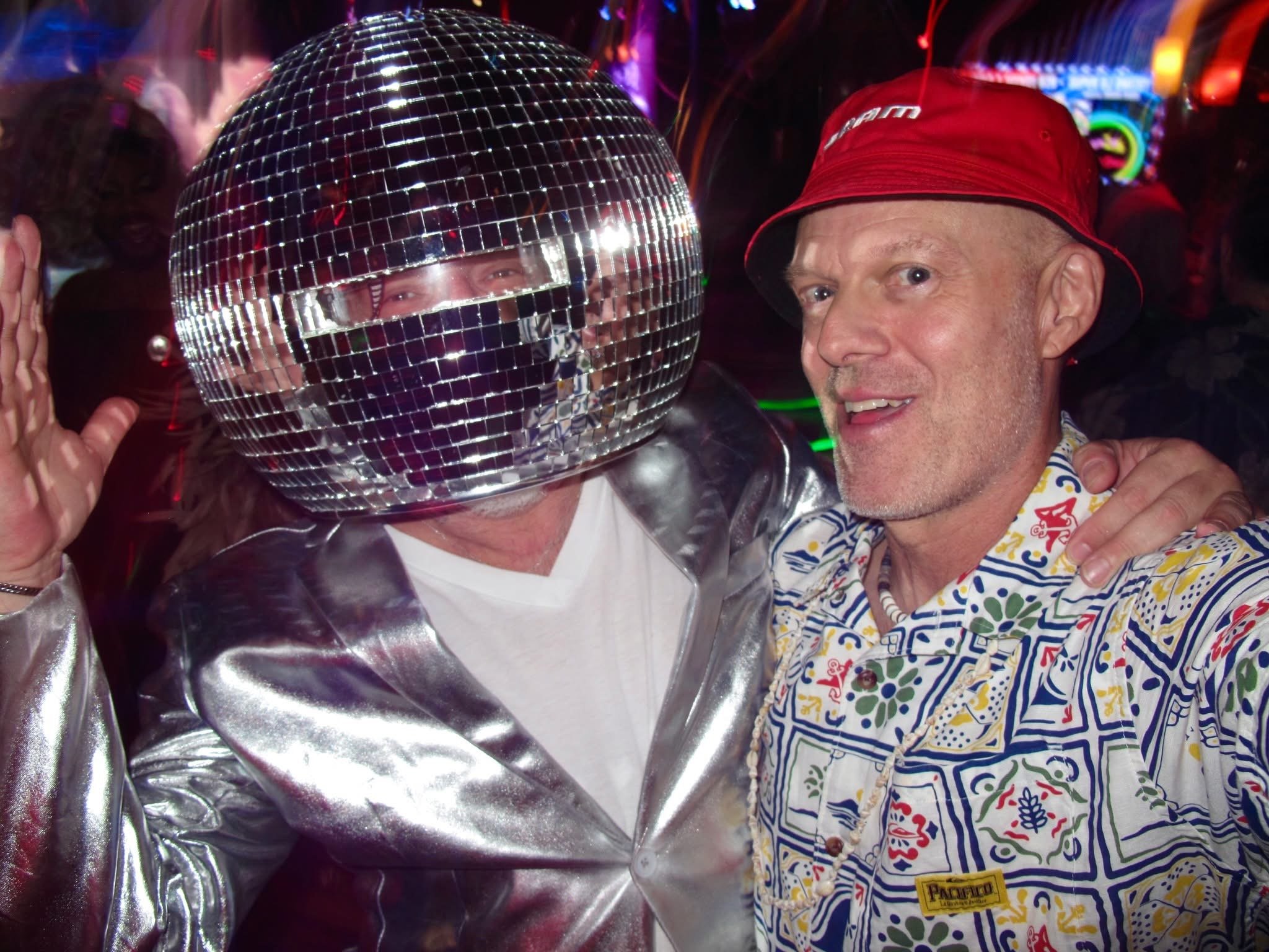 man wearing disco ball mask next to man in red hat and colorful button down shirt.