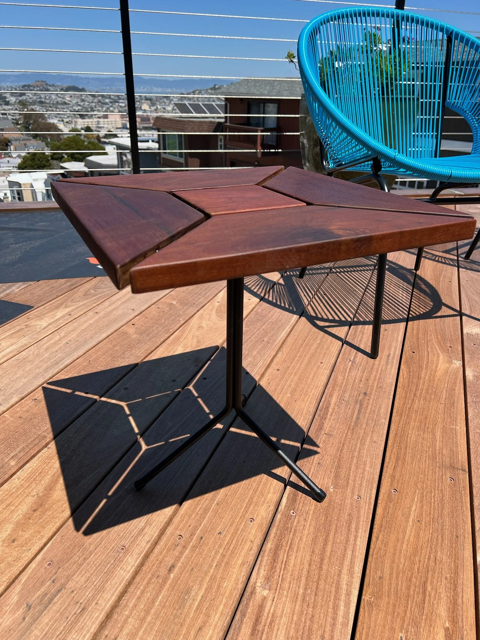 A wooden table with metal legs on a wooden deck, with a blue chair and a cityscape view with houses and mountains in the background.