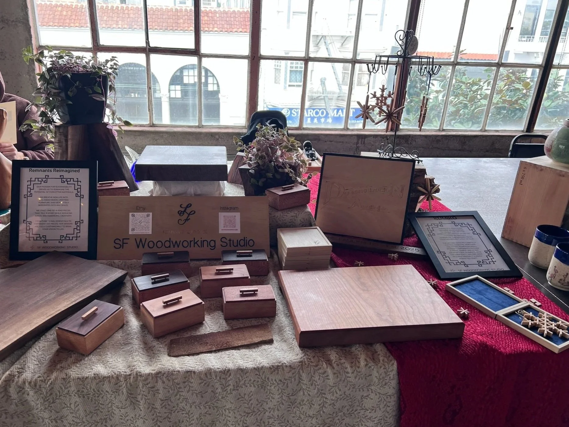 SF Woodworking Studio table display at The Box SF of handcrafted wooden items, signs, and framed documents, inside a room with large windows and plants in the background.