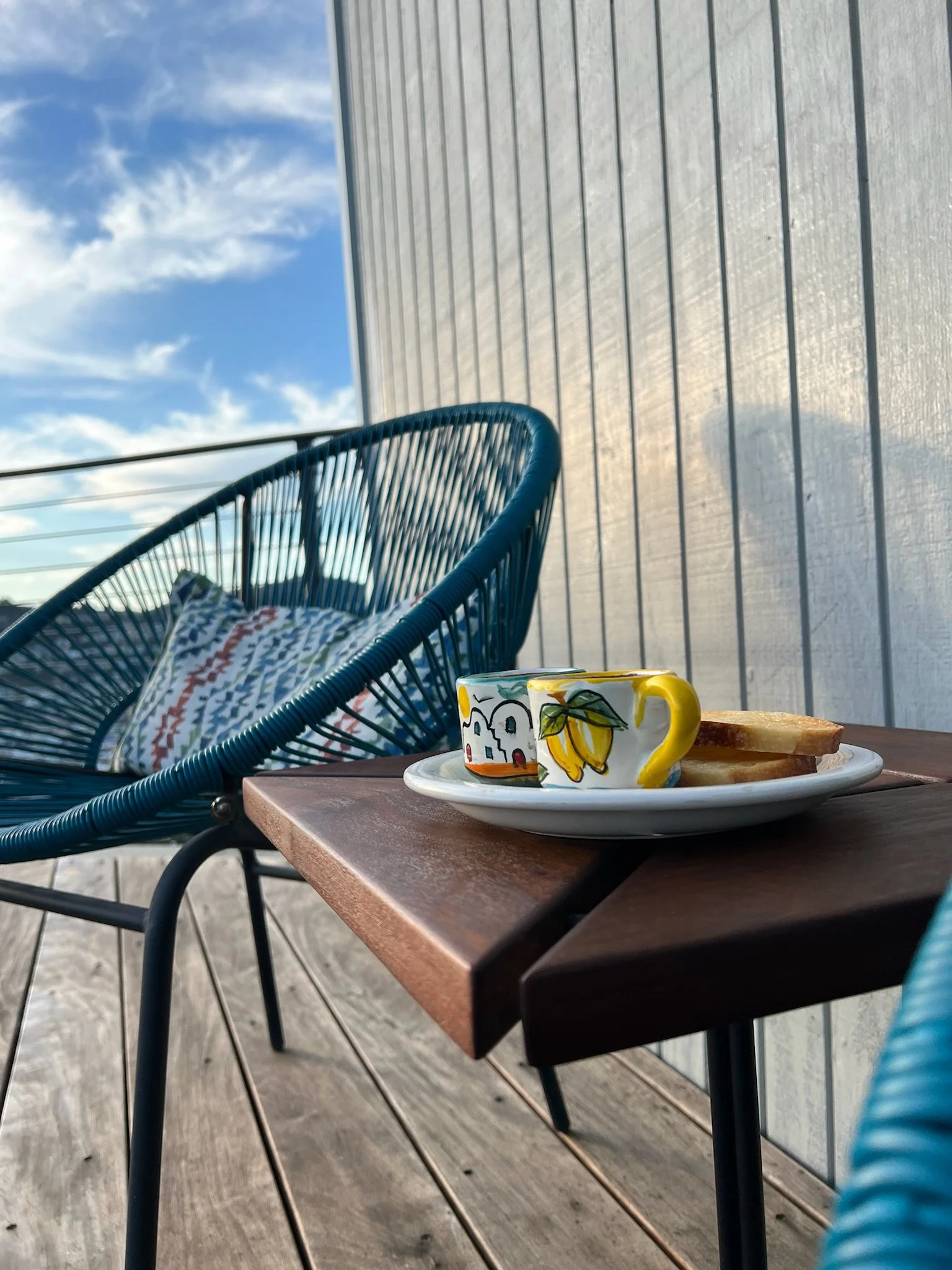 A small IPE wooden table with two colorful coffee mugs and some toast, on a balcony with blue woven chair and decorative pillow, under a partly cloudy sky.