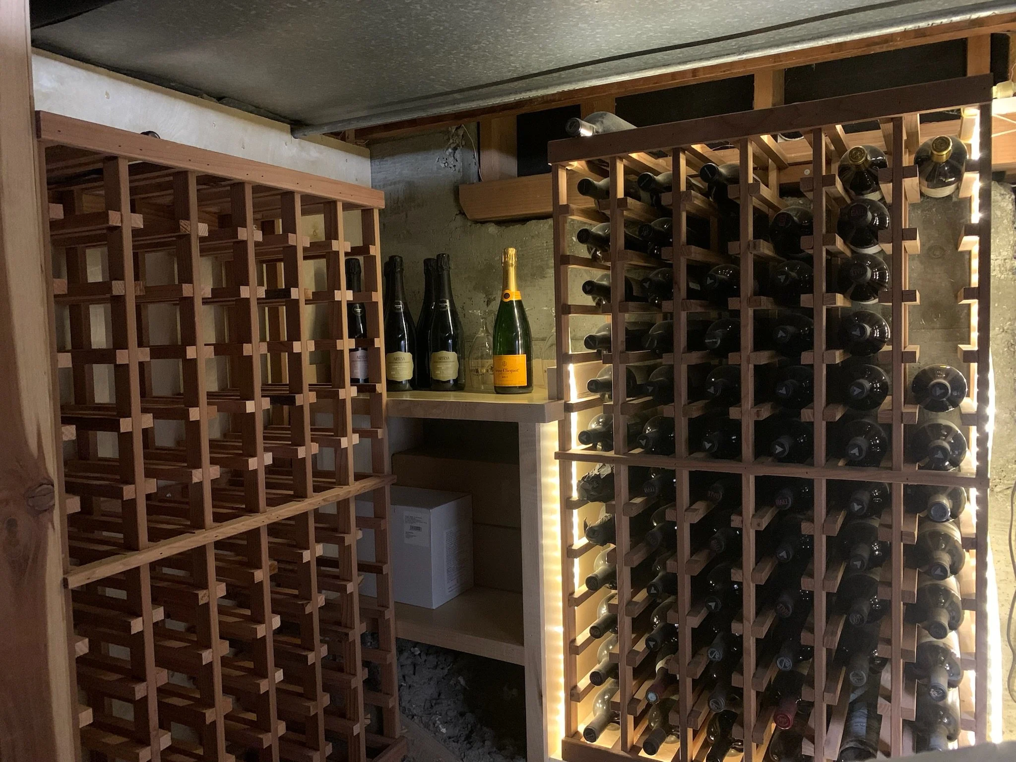 A wine cellar with wooden wine racks filled with wine bottles, some on the right side and some on the left side, and a small shelf in the middle holding three bottles of sparkling wine and a champagne bottle.