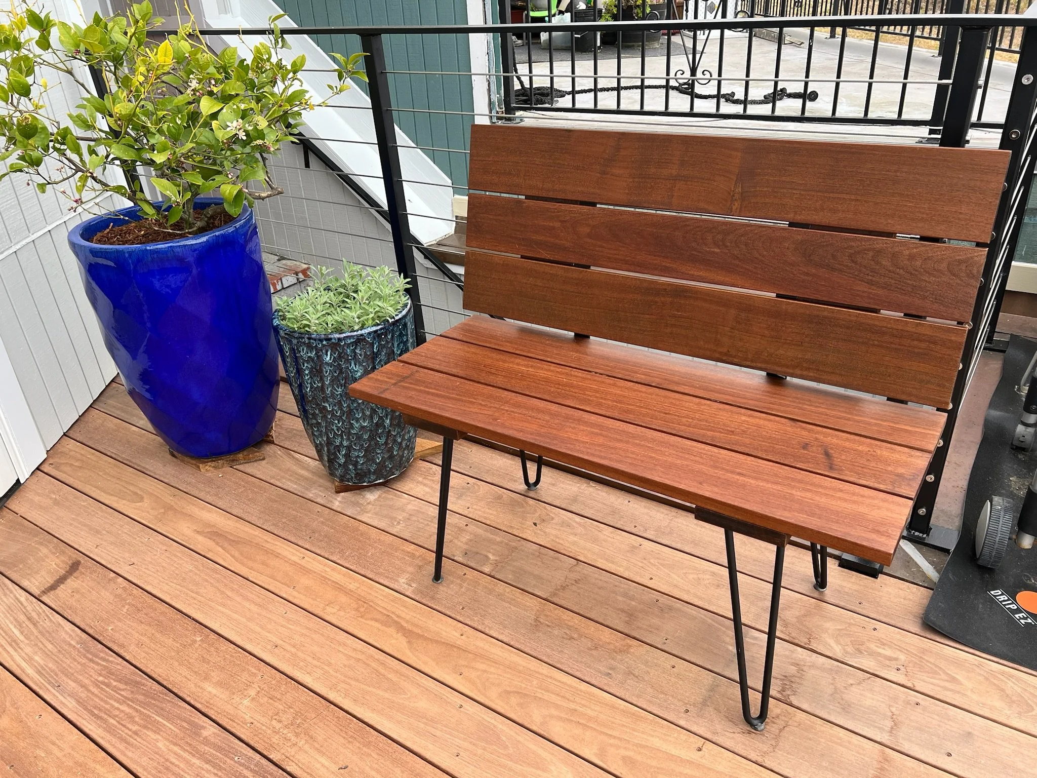 A patio with an IPE wooden bench and two ceramic planters, one large cobalt blue and one smaller with green foliage, on a wooden deck with a black metal railing.