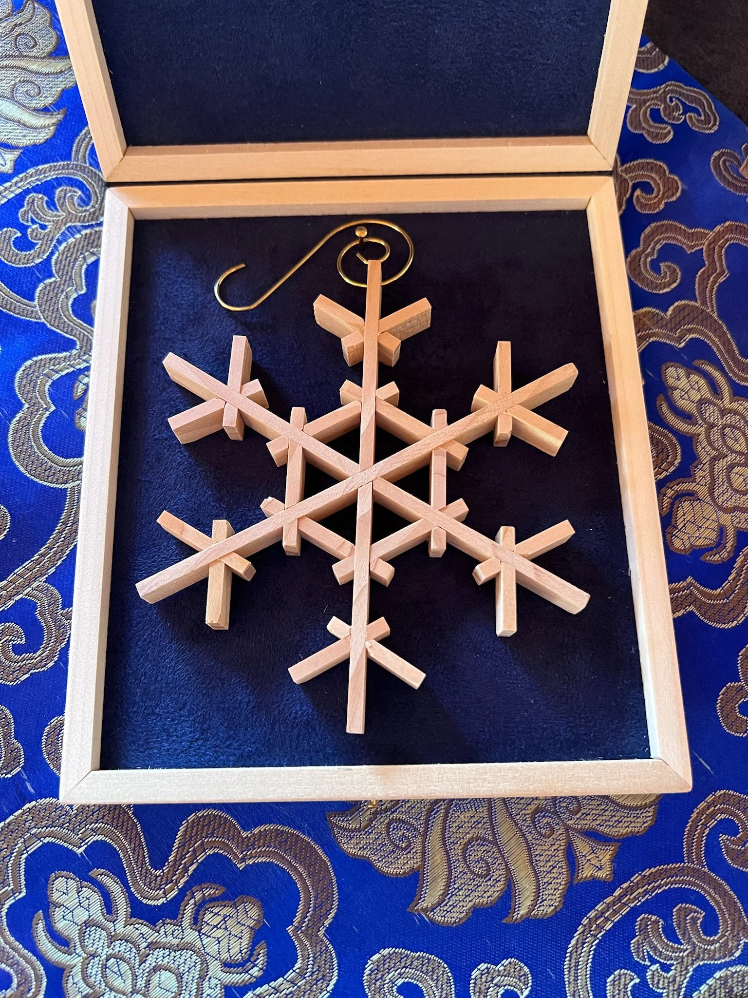 A wooden kumiko inspired snowflake ornament inside a wooden box with a blue suede interior, placed on a blue and gold patterned fabric.