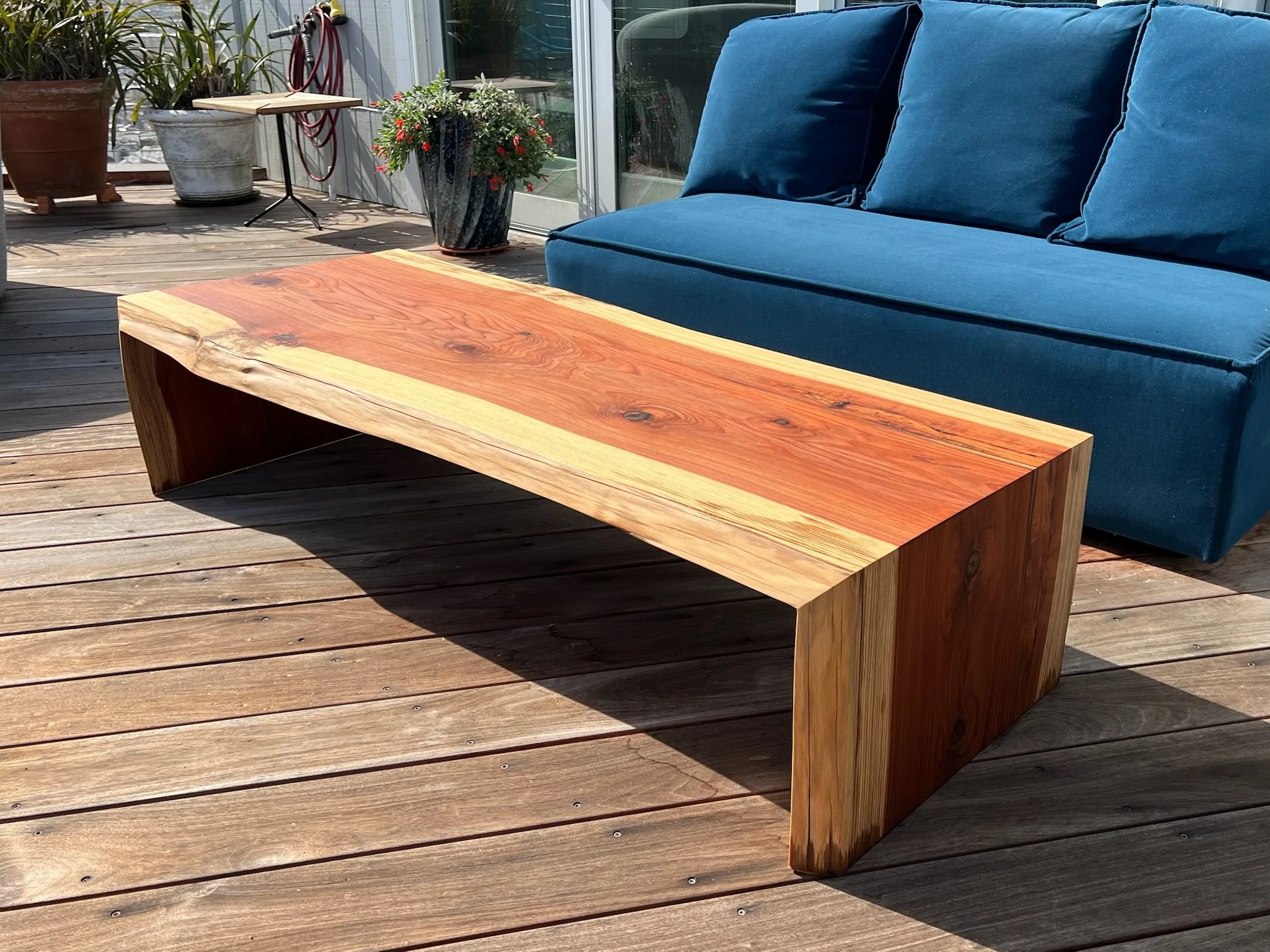 Redwood waterfall outdoor coffee table on a wooden deck with a blue outdoor sofa and potted plants in the background.