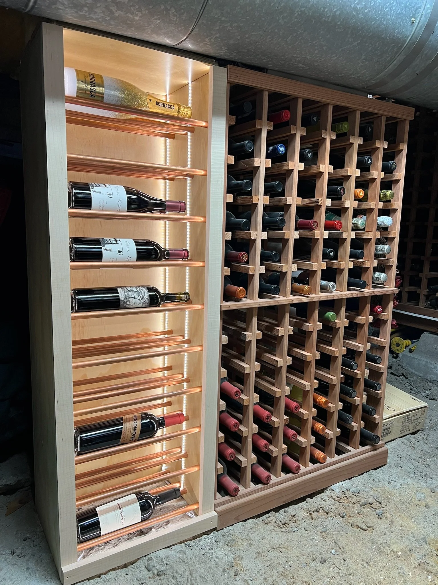 A wine rack with illuminated shelves on the left holding several bottles of wine and copper pipes, next to a wooden wine storage unit with multiple compartments filled with wine bottles.