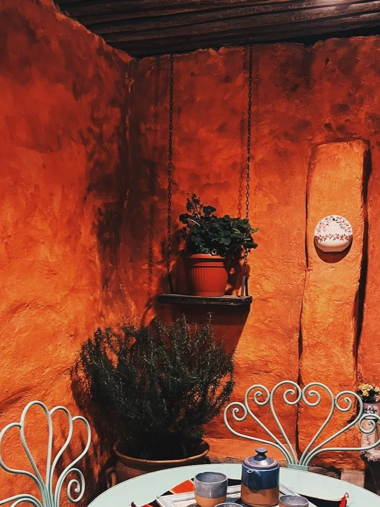Une terrasse intérieure avec un mur en terre cuite, une plante en pot suspendue, une autre plante en pot sur le sol, une table ronde avec des tasses et une théière en métal, et un décor vintage avec des chaises en métal peint en blanc. Argentine