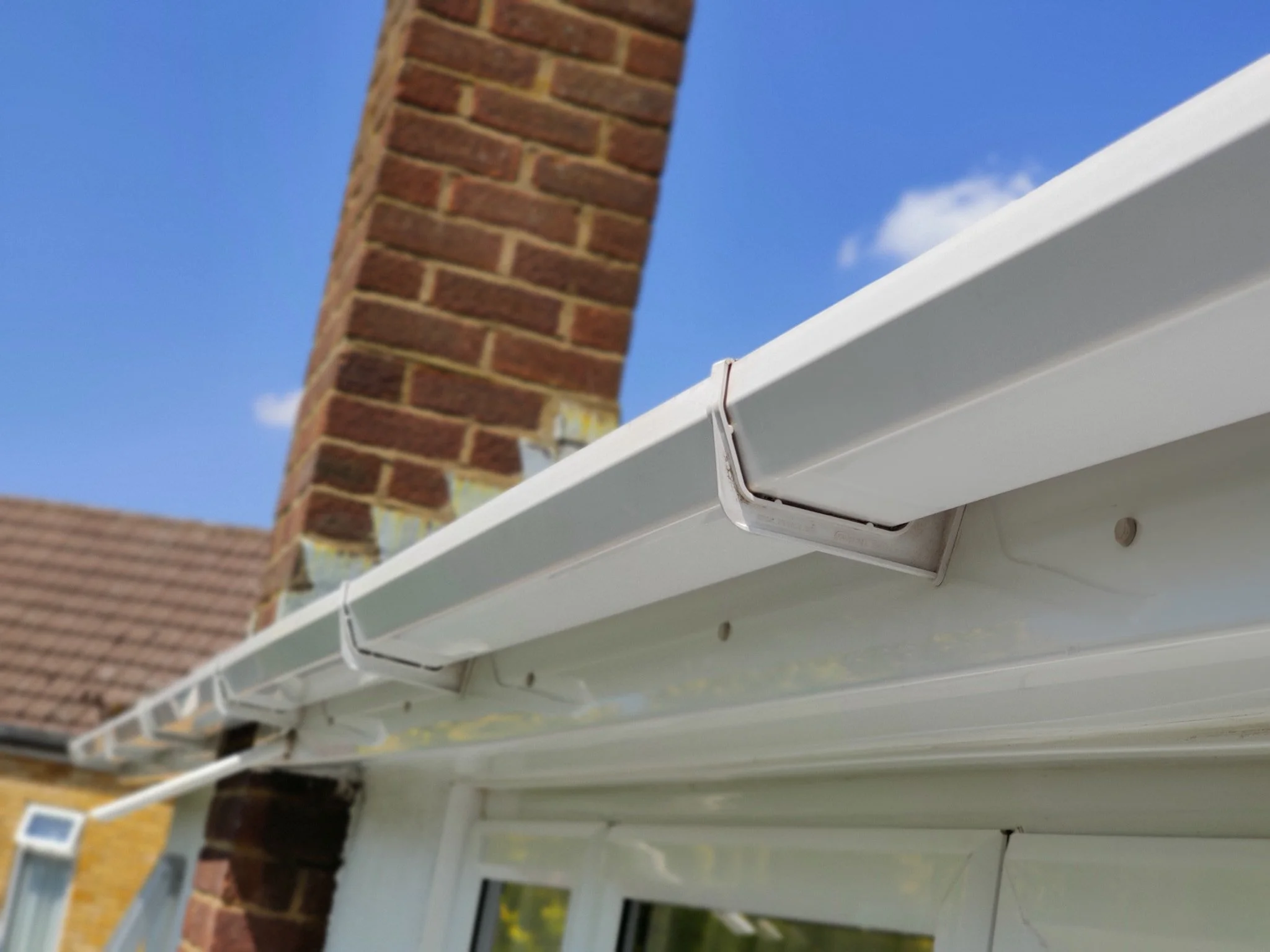 Close-up of a white gutter attached to a house roof with a brick chimney in the background and a blue sky with some clouds.
