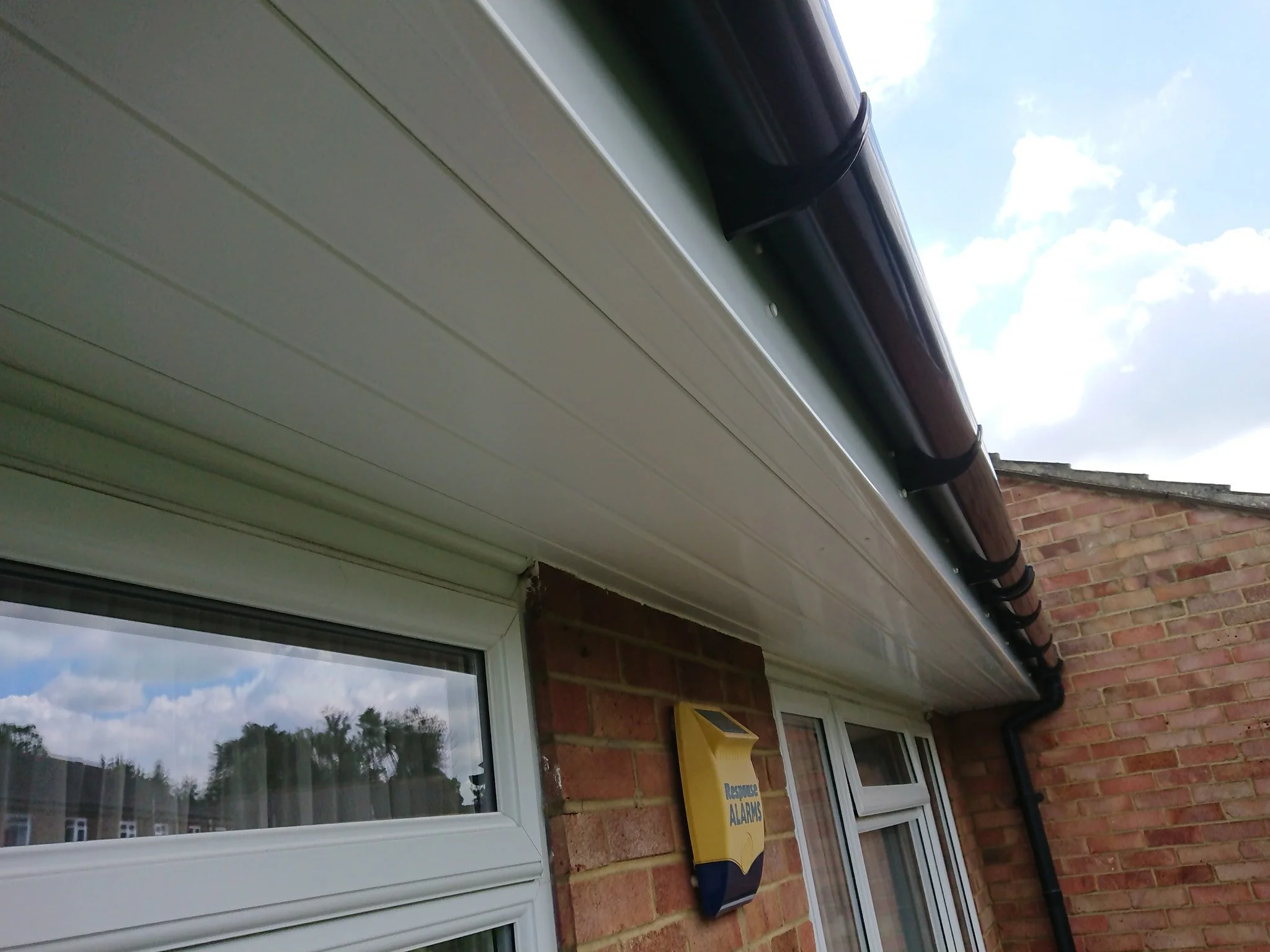 Close-up of the roof eaves of a brick house, showing a beige gutter and downspout system against a background of partly cloudy sky.