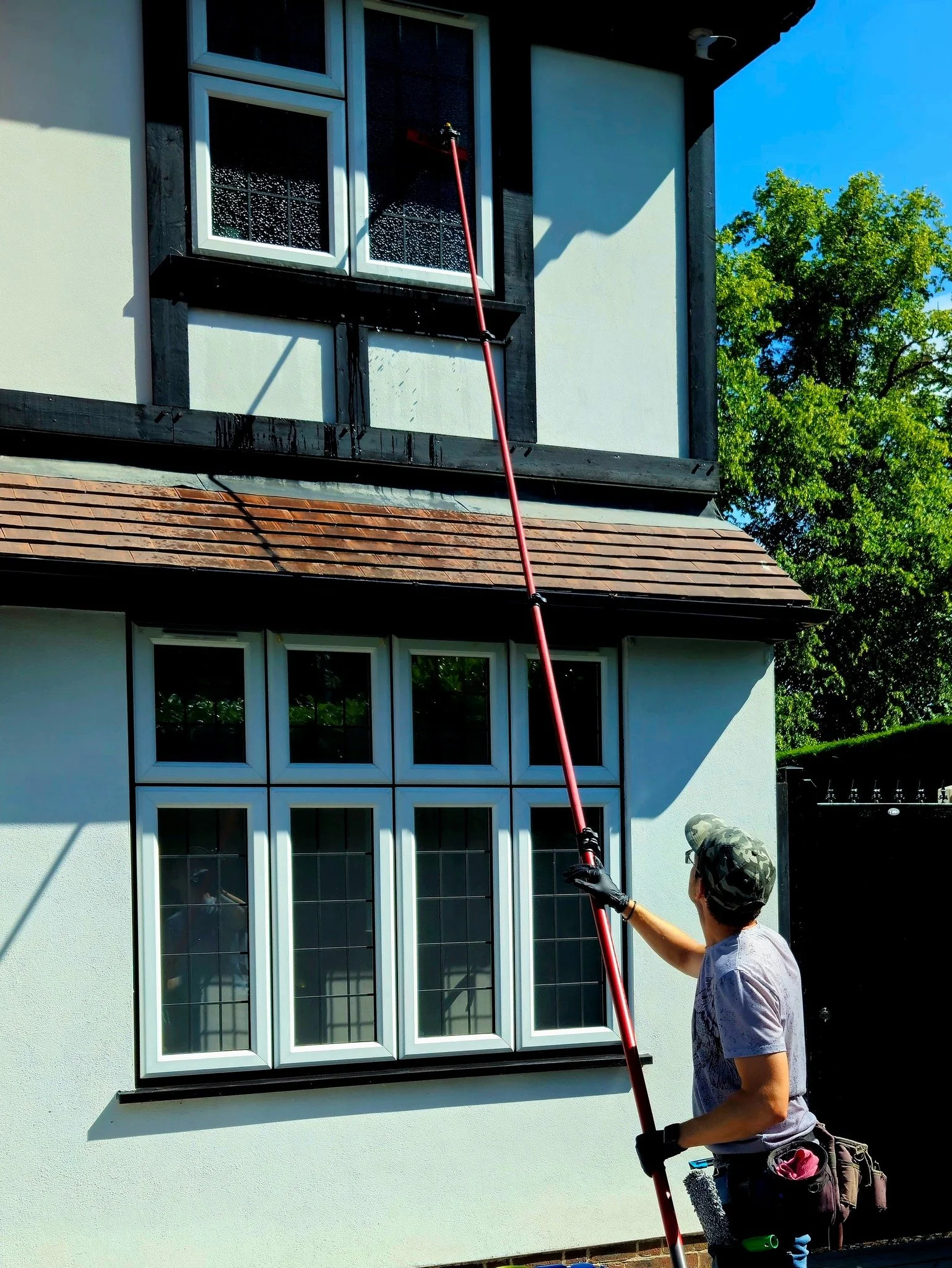 A person cleaning the windows of a two-story house using an extendable cleaning tool with a water spray. The house has white walls, multiple windows, and a brown tiled roof, with trees visible in the background.
