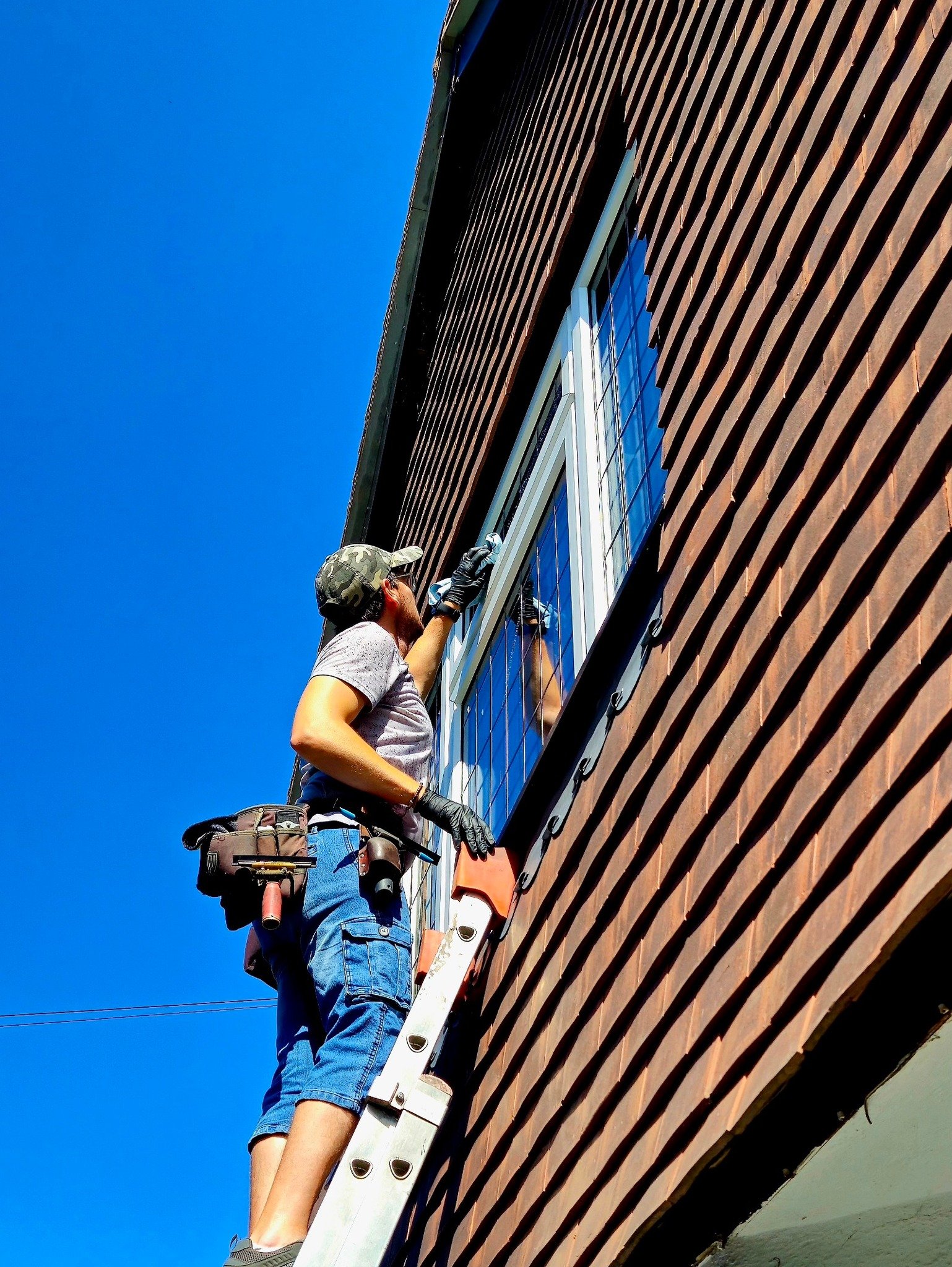 A man in casual clothes, wearing a camouflage hat and utility belt, is standing on a ladder cleaning or inspecting the windows of a building with dark brown siding on a clear day.